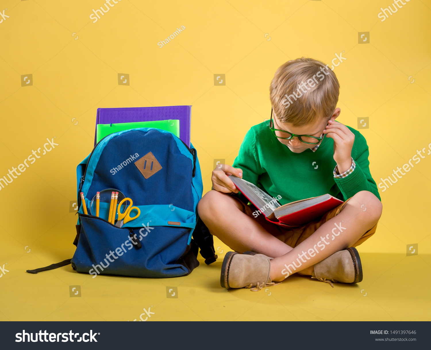 Children go back to school. Stylish boy doing homework at home with backpack full of books, pencils. Pupil reading a book, writing and painting. Kid is drawing. Child in glasses on yellow wall. #1491397646