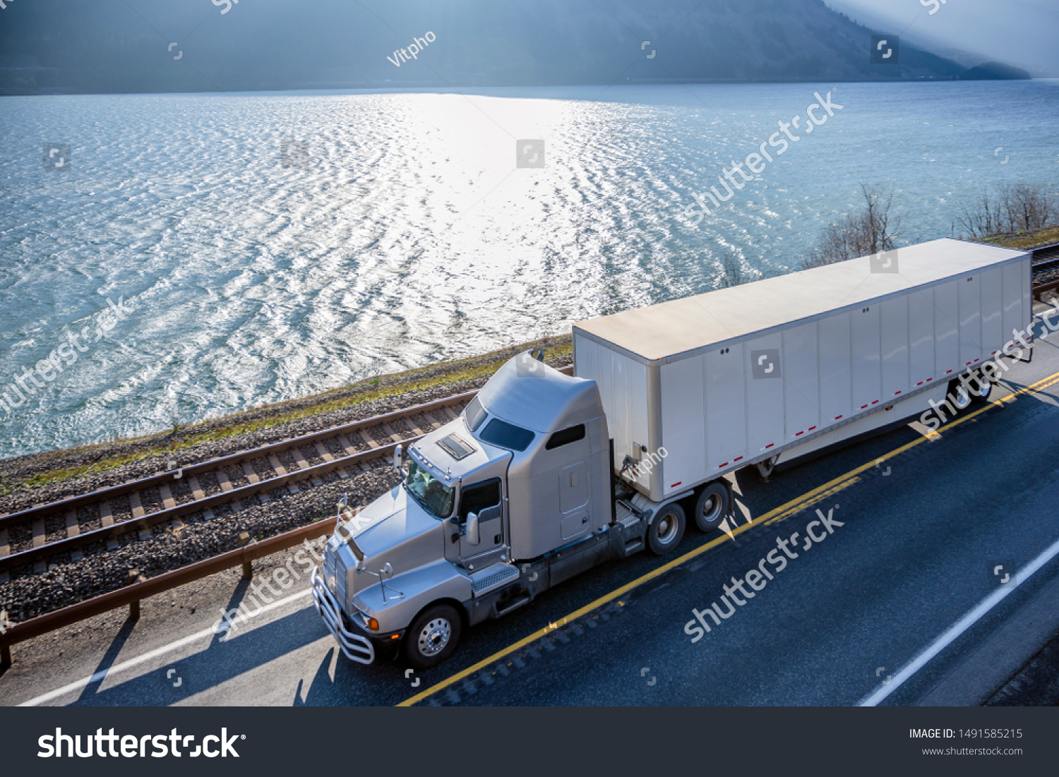 Big rig classic American powerful gray semi truck with dry van semi trailer transporting commercial cargo running on the road along railroad and river in Columbia Gorge area with mountain ranges #1491585215