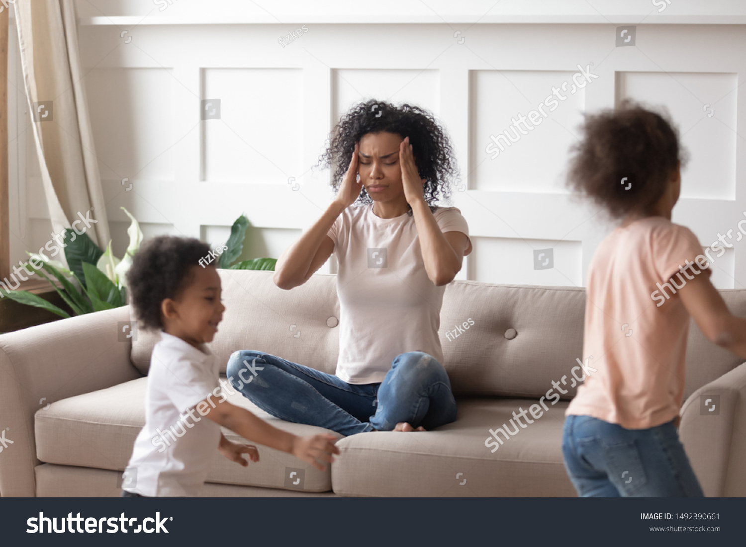 African mother sit on sofa closed eyes touch temples having severe headache or migraine because of noise of little crazy children running near her. Tired exhausted mom, upbringing difficulties concept #1492390661