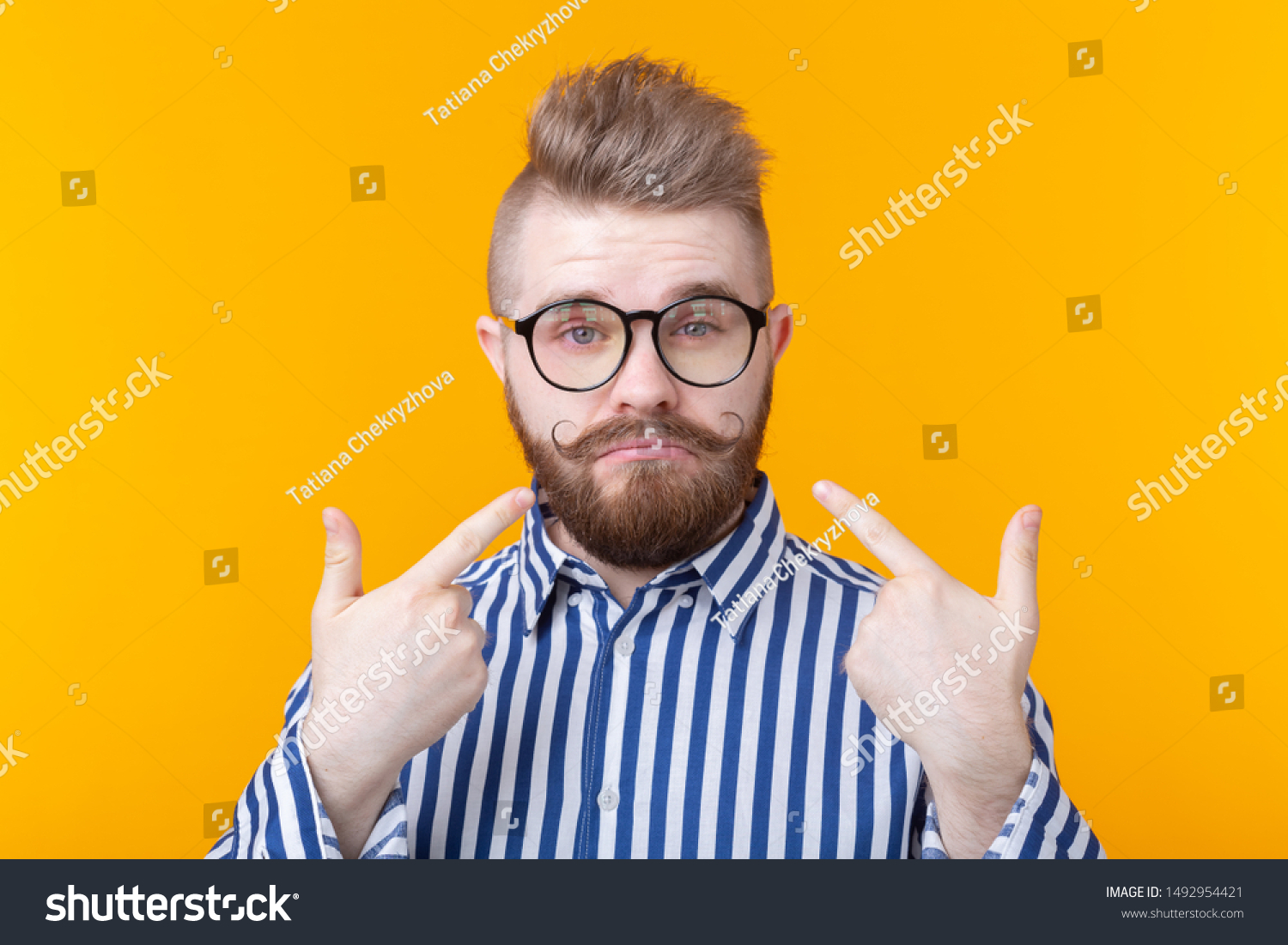 Charming confident young fashion hipster man with glasses and a beard shows on himself posing over a yellow background. Place for advertising. The concept of self-confidence and success. #1492954421