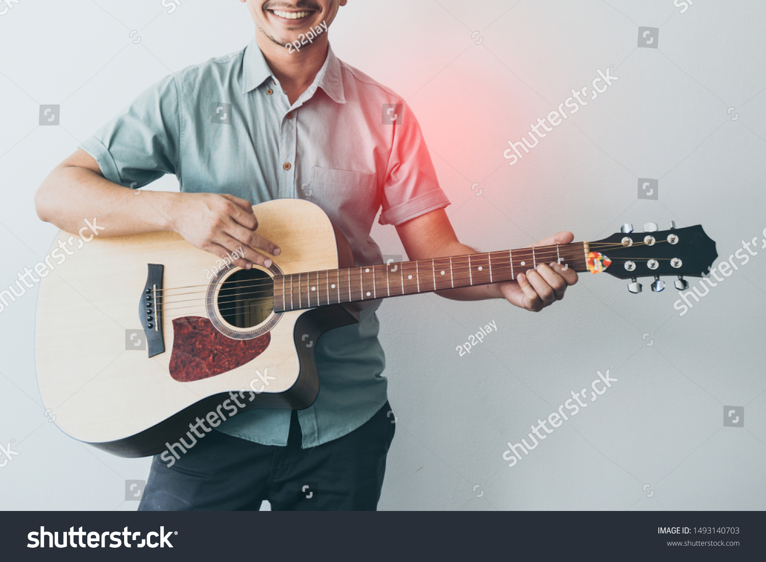 Cheerful guitarist. Cheerful handsome young man playing guitar and smiling while standing on white wall background, process color. #1493140703