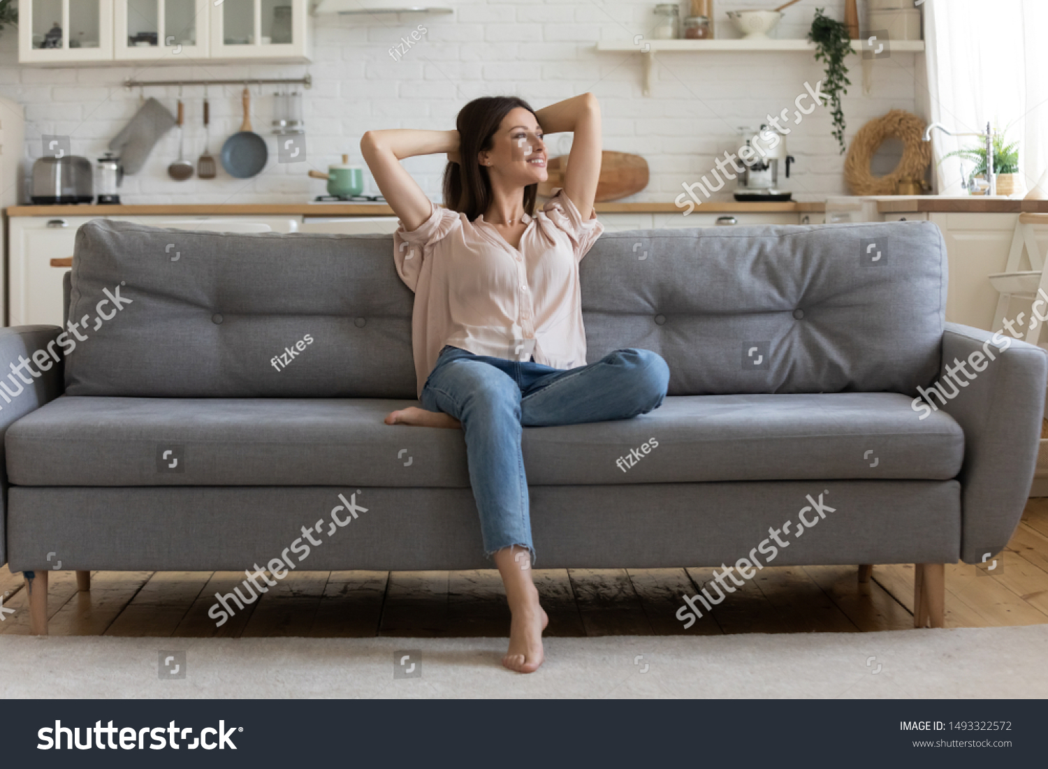 In cozy living room happy woman put hands behind head sitting leaned on couch 30s european female enjoy lazy weekend or vacation, housewife relaxing feels satisfied accomplish chores housework concept #1493322572