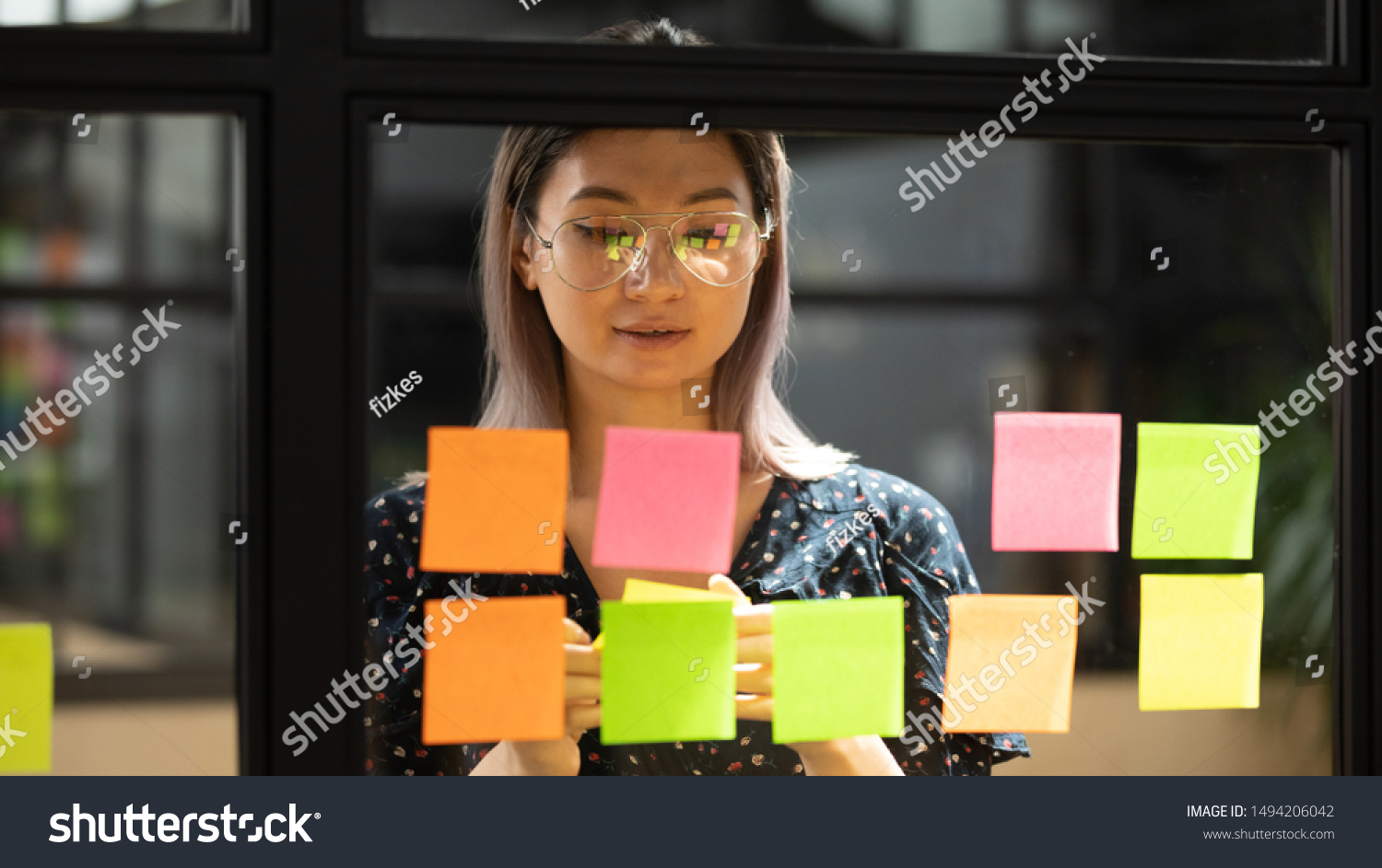 Serious focused young female coach teacher student asian business woman working on project strategy plan writing target tasks creative ideas on sticky post it notes on glass scrum board office wall #1494206042