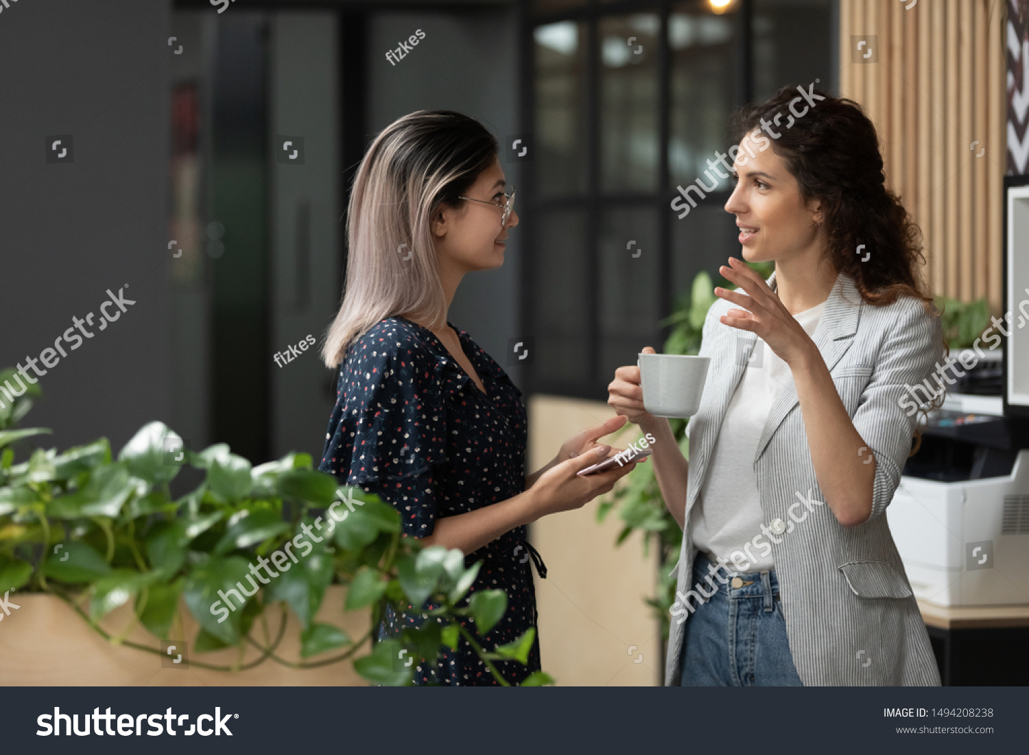 Two young diverse business women chatting during work break standing in modern office, caucasian female employee share ideas talking with asian colleague discussing team project at corporate meeting #1494208238