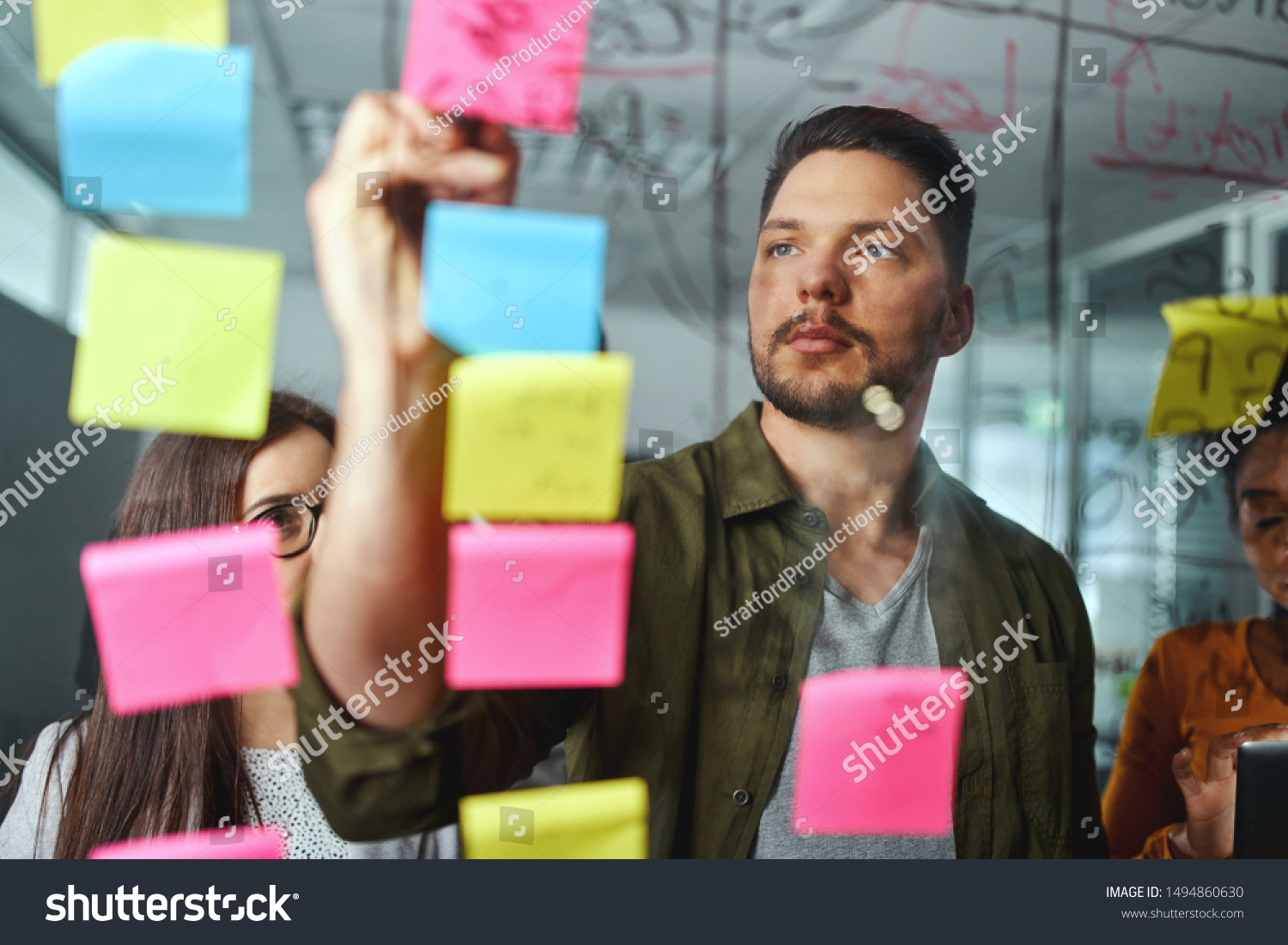 Concentrated male executive discussing business ideas with colleagues by writing on sticky notes pasted over the glass board #1494860630