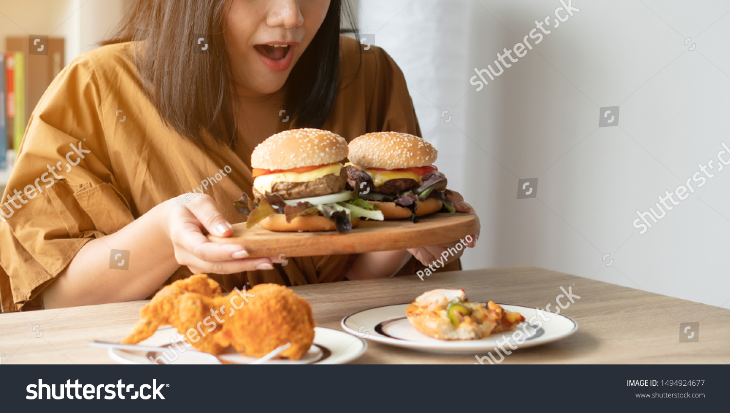 Hungry overweight woman holding hamburger on wooden plate, Fried chicken and Pizza on table .Concept of binge eating disorder (BED). #1494924677