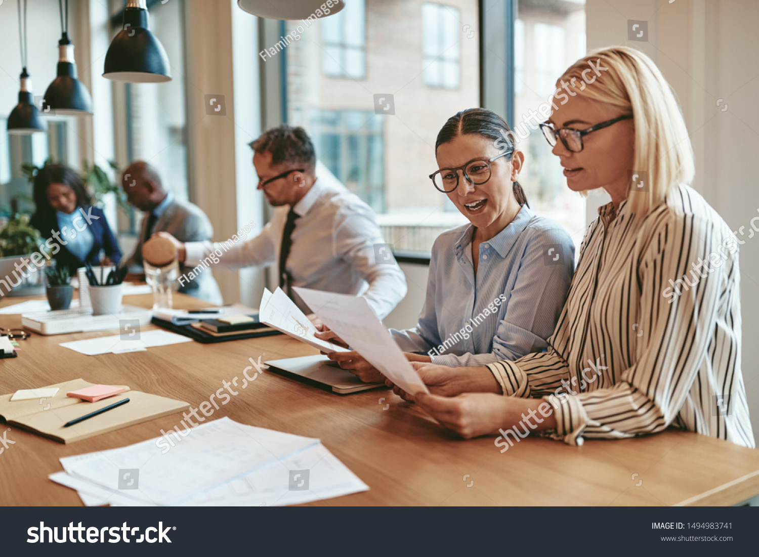Two smiling young businesswomen reading paperwork together while sitting at a boardroom table during an office meeting #1494983741