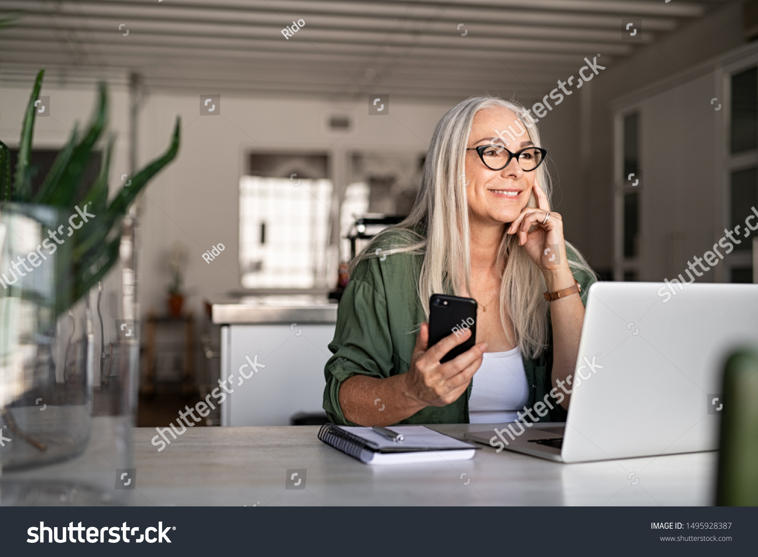 Happy senior woman holding smartphone and laptop daydreaming while looking away. Successful stylish old woman working at home while thinking. Fashionable lady entrepreneur wearing cool eyeglasses. #1495928387