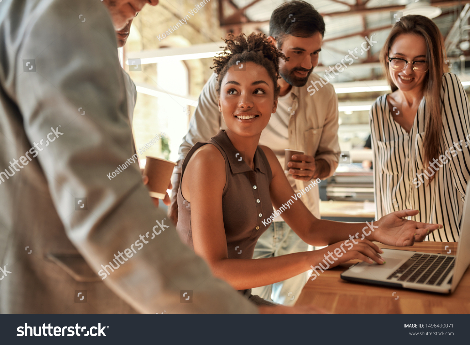 Working together. Young and cheerful afro american woman using laptop and discussing something with colleagues while sitting in the modern office. Teamwork. Office life #1496490071