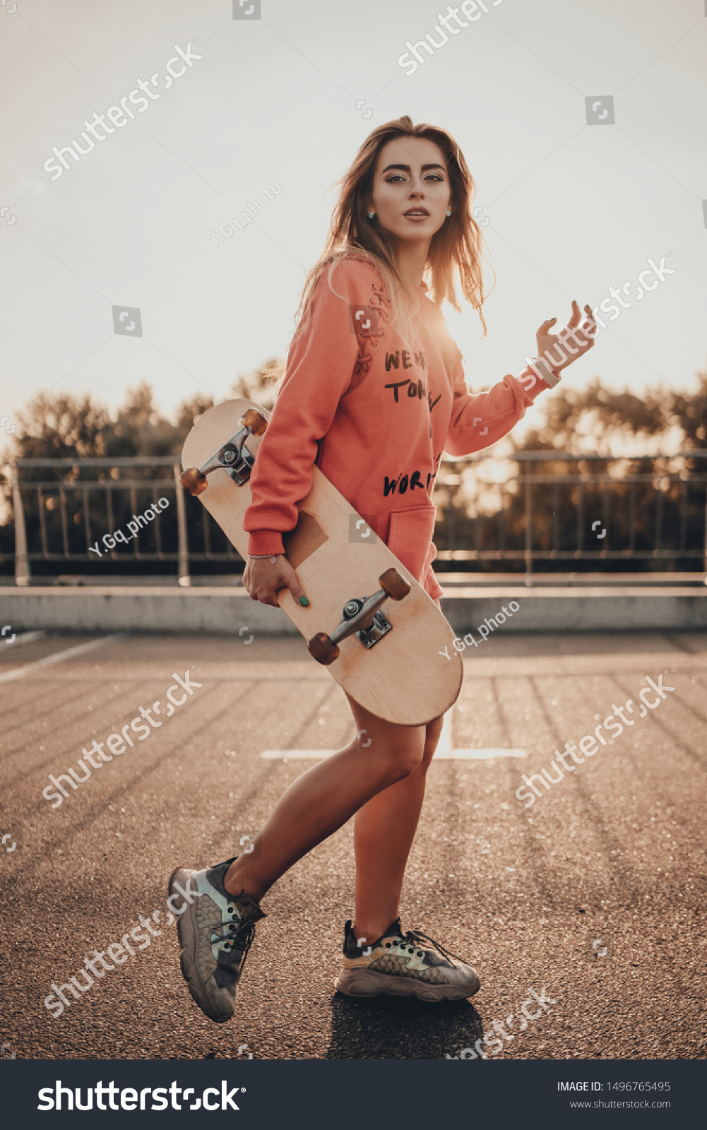 Urban stylish young girl walking with skateboard in red hoodie and sneakers on summer sunset on parking near road. Outdoors portrait of active sport woman model #1496765495