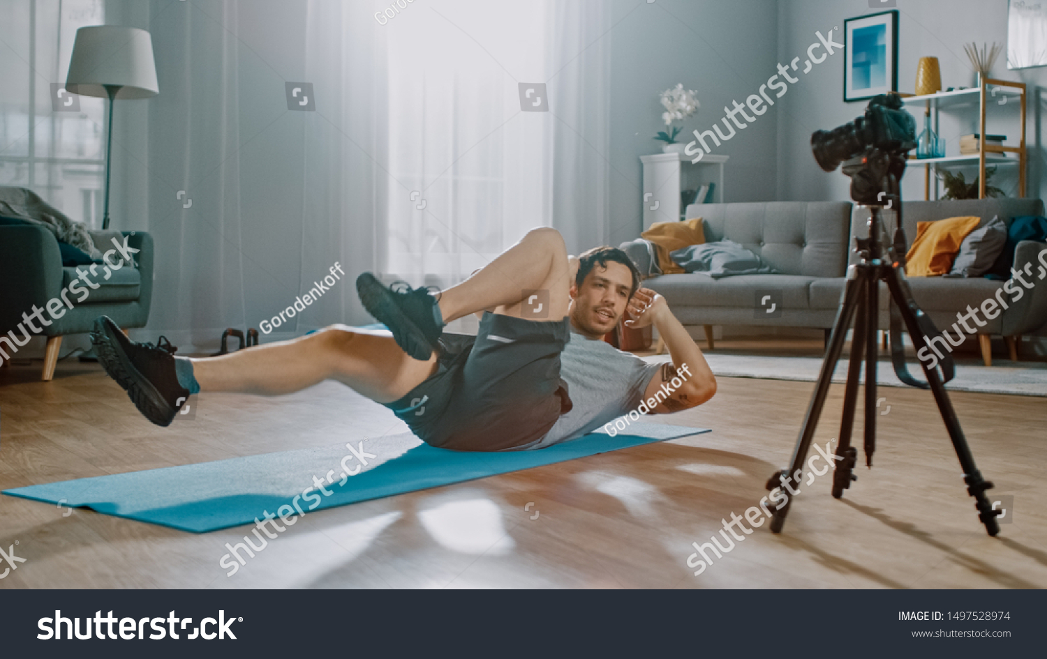 Strong Athletic Fit Man in T-shirt and Shorts is Recording his Crisscross Crunch Workout on Camera for His Blog. Scene takes place in His Spacious and Bright Living Room with Minimalistic Interior. #1497528974