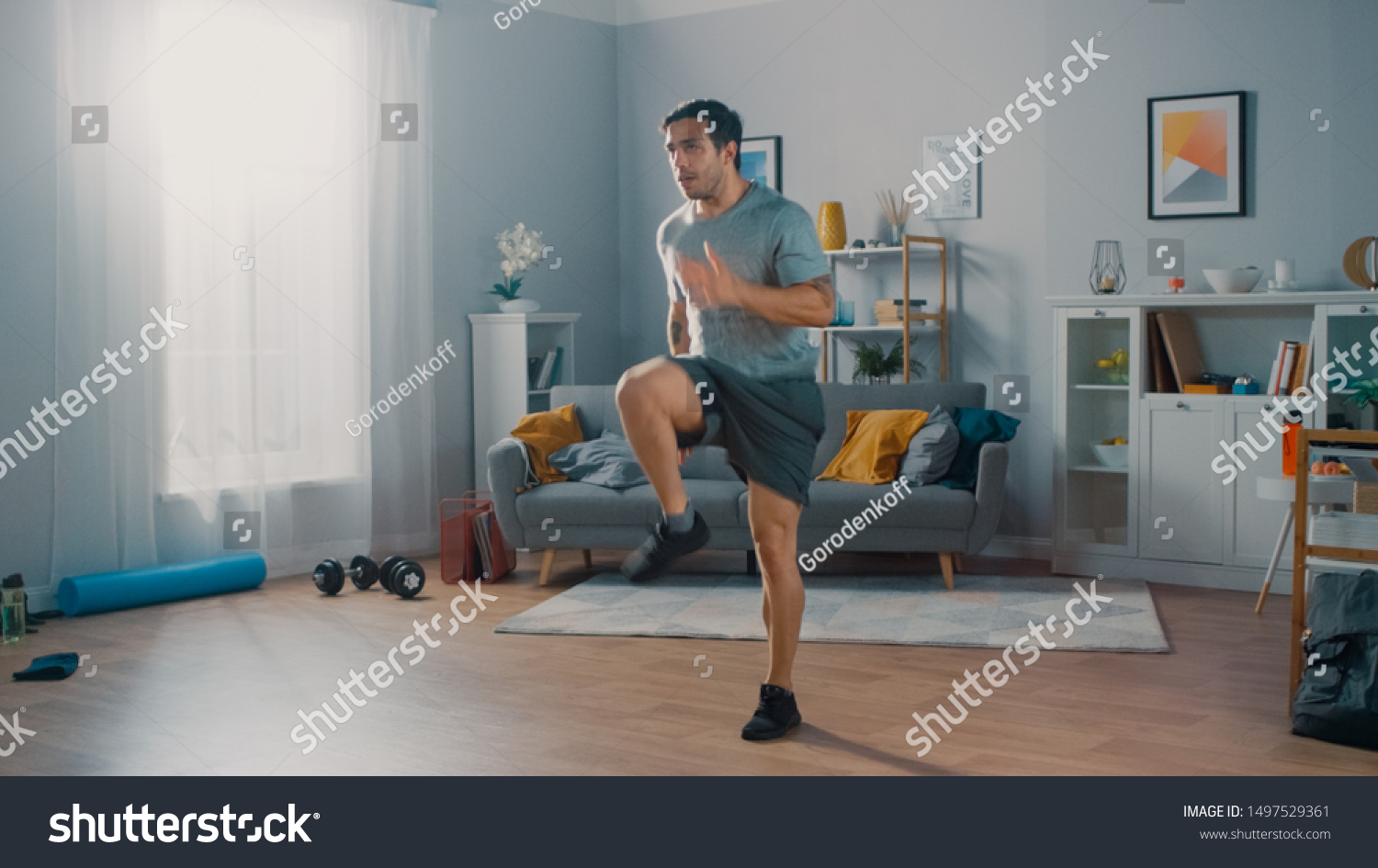 Strong Athletic Fit Man in T-shirt and Shorts is Energetically Jogging in Place at Home in His Spacious and Bright Living Room with Minimalistic Interior. #1497529361