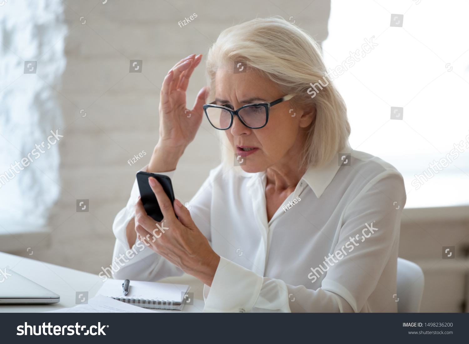 Confused senior businesswoman sit at office desk hold cellphone experience internet connection problem, frustrated aged woman worker feel disappointed having smartphone breakdown or virus attack #1498236200