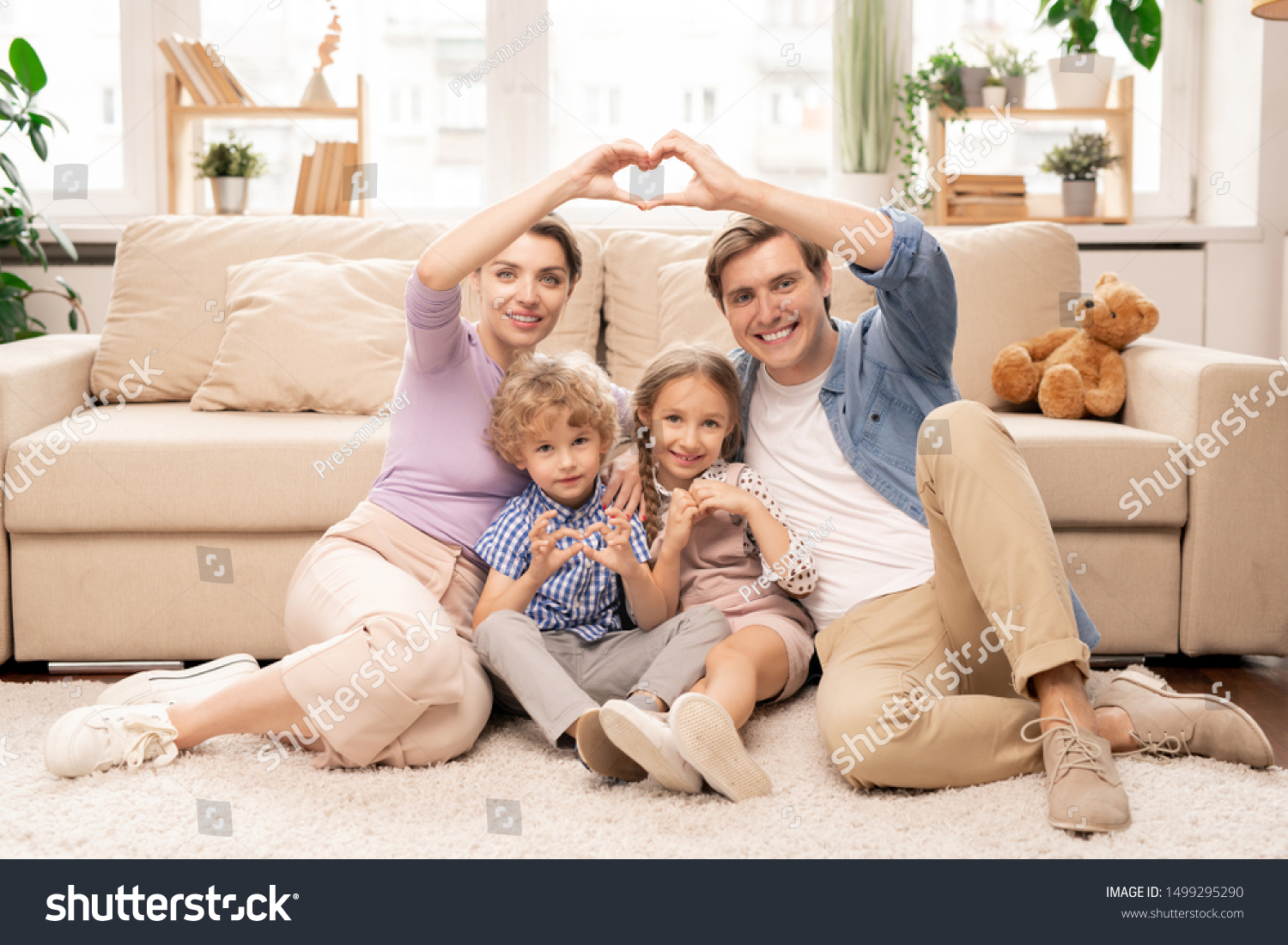 Happy parents and two adorable siblings sitting on the floor by couch in front of camera and making heart shape with fingers #1499295290