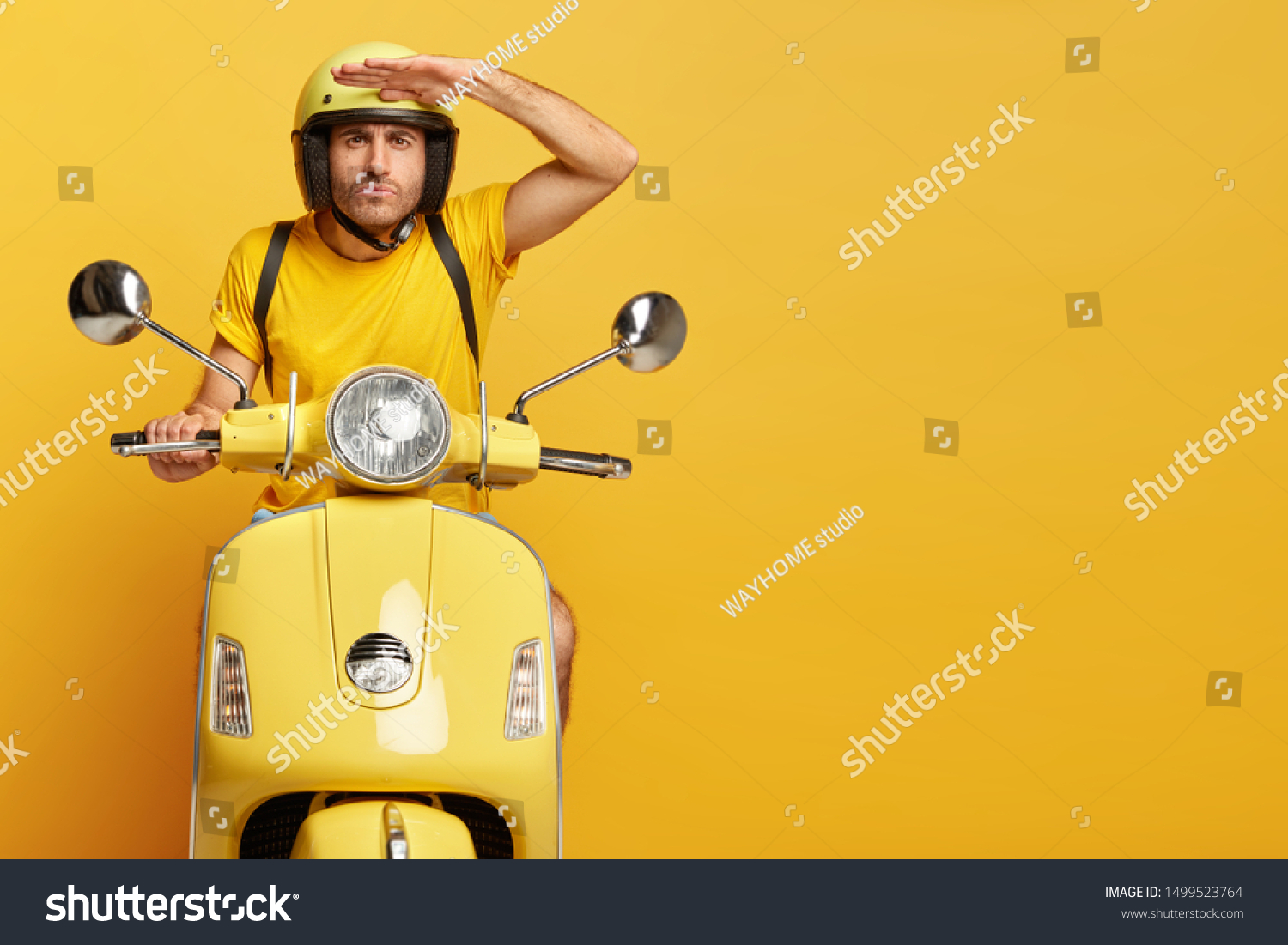 Concentrated male driver in helmet on scooter, keeps palm near forehead, poses on motorbike, carries rucksack, has serious face expression, poses against yellow background, copy space on right side #1499523764