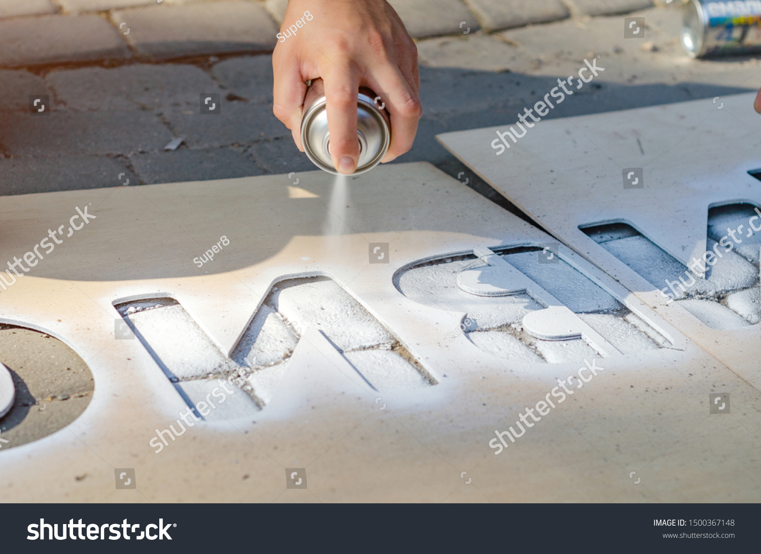 Hand holds an aerosol spray can, puts the image on the asphalt through a stencil #1500367148