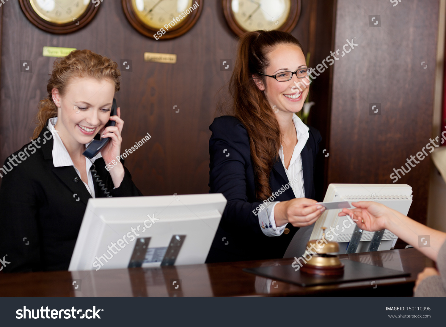 Two beautiful young stylish receptionists at a reception desk, one talking on the telephone and the other handing a card to a customer #150110996