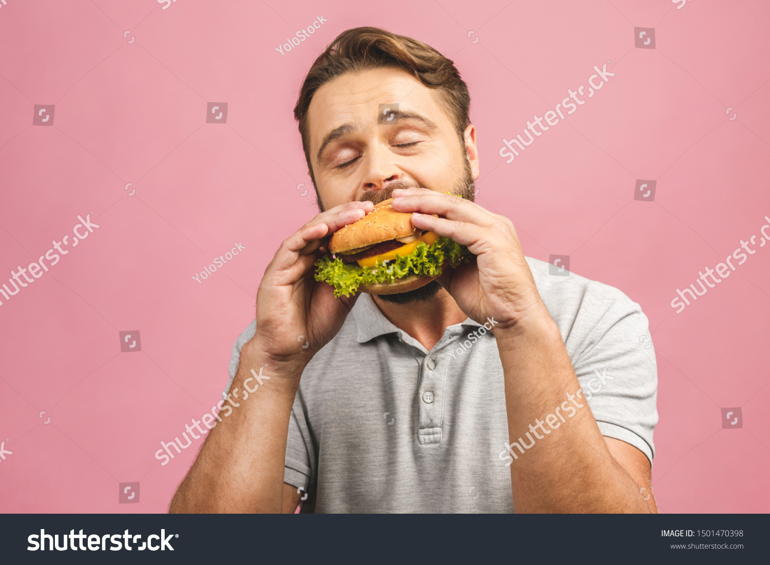 Young man holding a piece of hamburger. Bearded gyu eats fast food. Burger is not helpful food. Very hungry guy. Diet concept. Isolated over pink background.