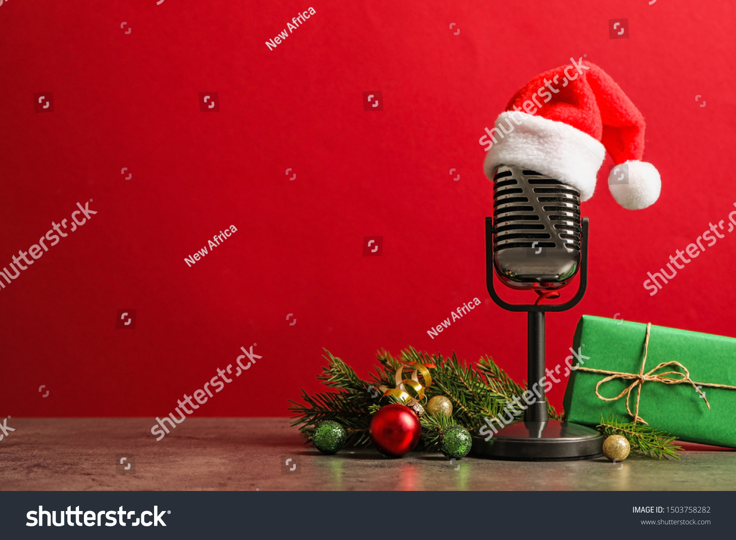 Microphone with Santa hat and decorations on grey table against red background, space for text. Christmas music #1503758282