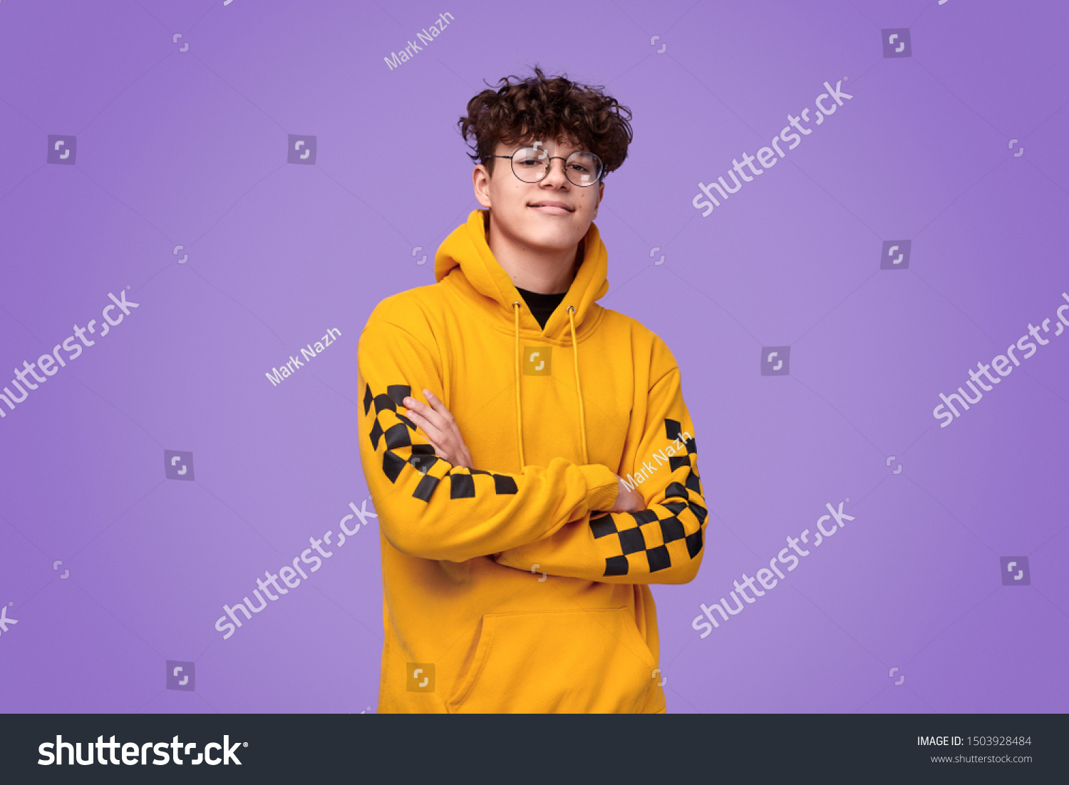 Positive teen boy in yellow hoodie crossing arms and looking at camera against bright violet background #1503928484