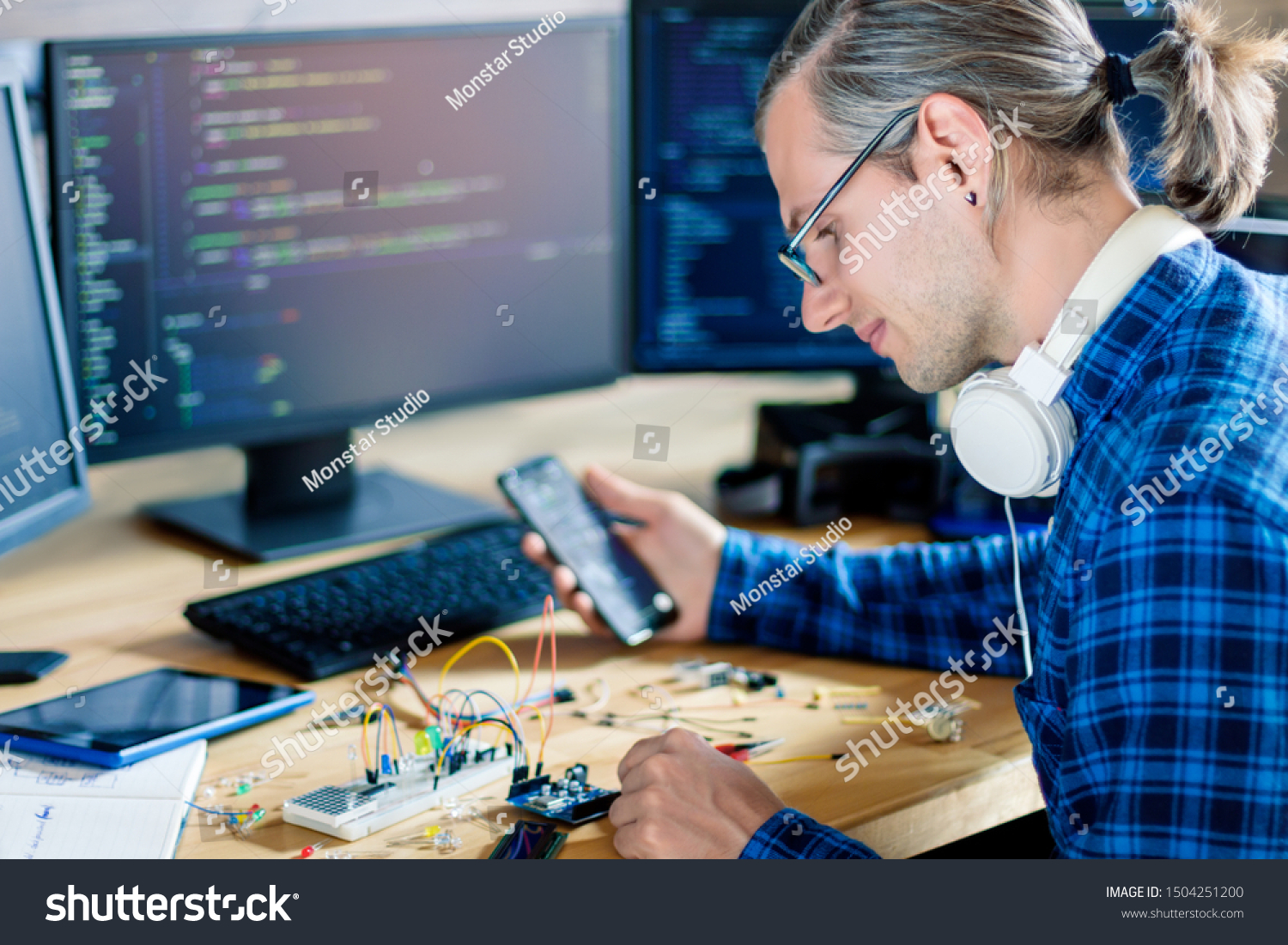Developer is connecting breadboard to microcontroller. Man is holding smartphone with program code software for controlling electronic device. Chips, resistors, diodes on desktop of hardware engineer. #1504251200