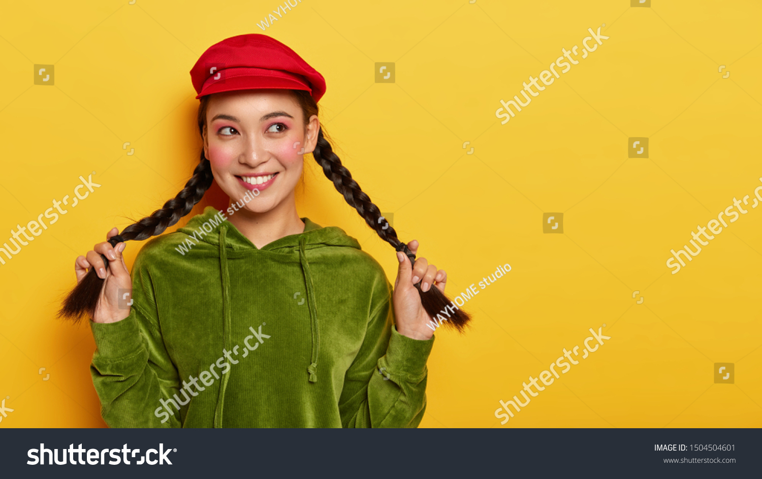Good looking brunette woman holds two pigtails, looks away with happy expression, thinks about interesting choice, recalls lovely moment in life, wears red cap and green jumper, smiles pleasantly #1504504601
