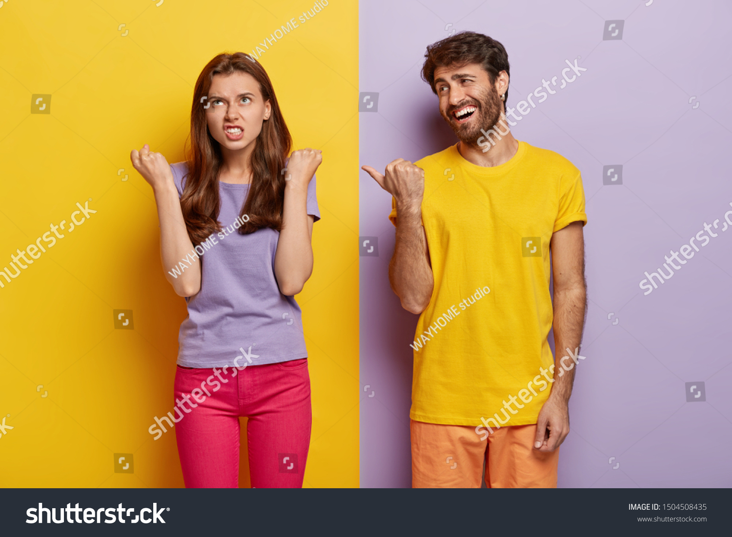 Pleased guy has fun, wears bright yellow t shirt, points thumb at irritated girlfriend who clenches fists with anger, expresses negative emotions, stand in studio against colorful background. Feelings #1504508435
