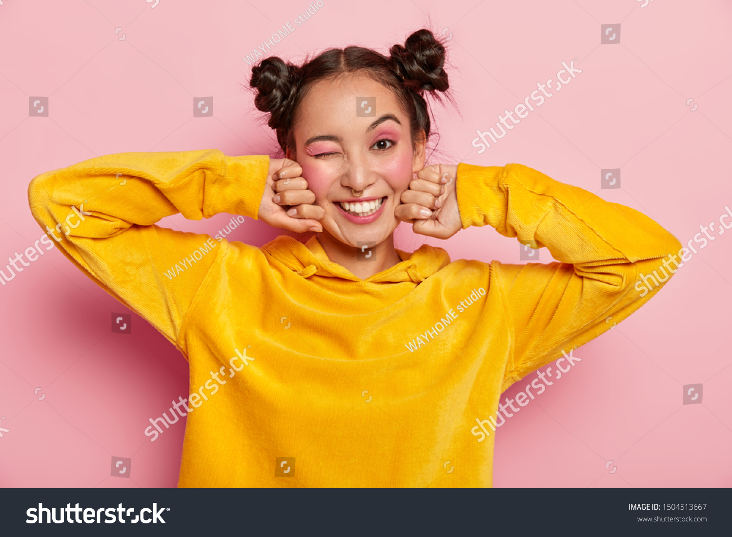 Image of lovely young Asian woman with happy face expression, blinks eye and smiles positively, has fun indoor, two hair knots, dressed in yellow velvet hoody, isolated over pink background. #1504513667
