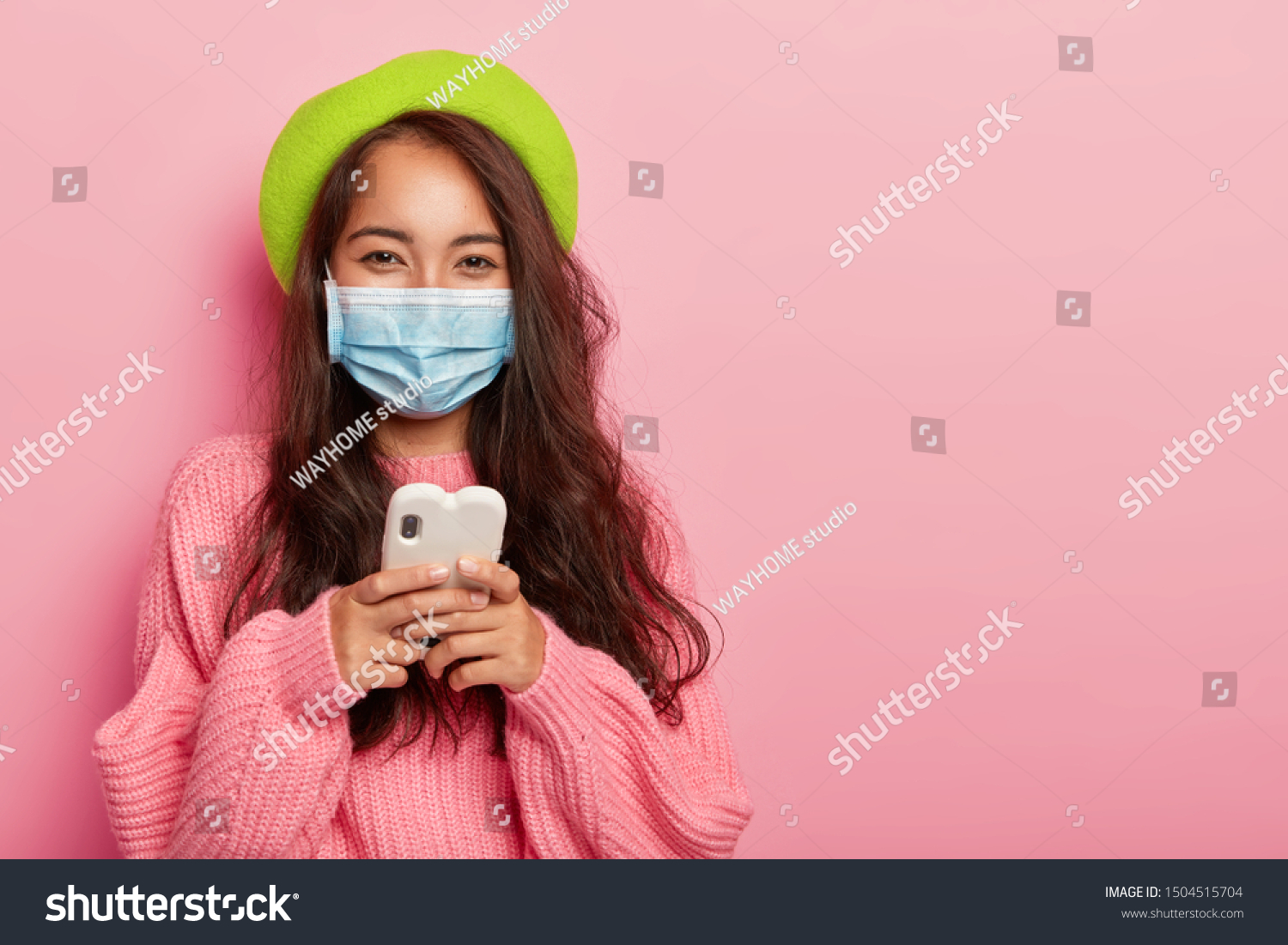 Satisfied mixed race woman has flu, wears protective medical mask not to infect other people, uses cell phone for surfing internet, reads how to cure disease online, dressed in fashionable clothes #1504515704