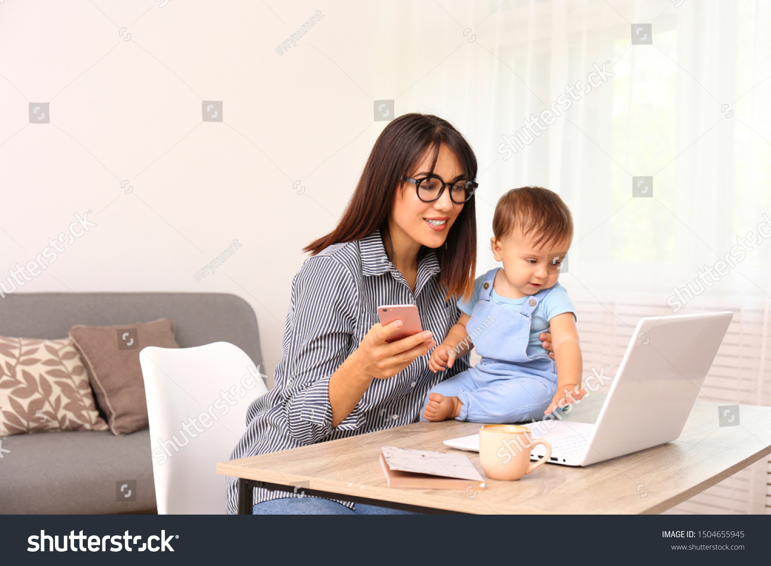 Stay at home mom working remotely on laptop while taking care of her baby. Young mother on maternity leave trying to freelance by the desk with toddler child. Close up, copy space, background. #1504655945