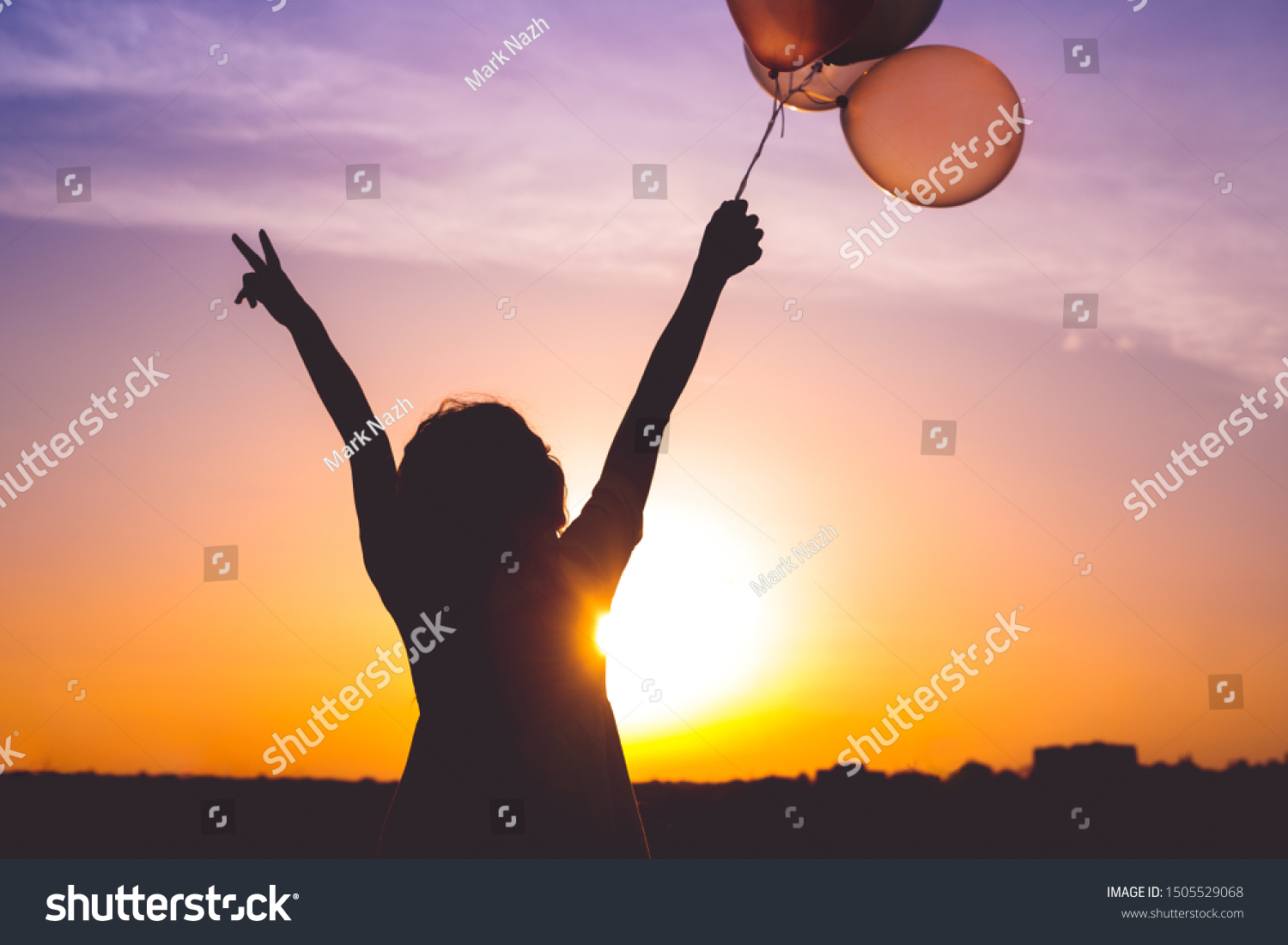Silhouette of female holding bunch of balloons in raised arms and gesturing V sign against bright sundown sky #1505529068