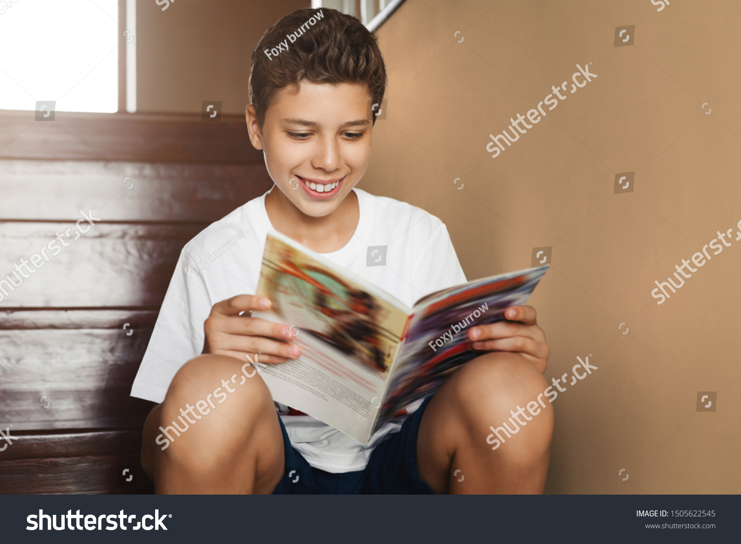 Smiling attractive teen boy sitting at home on steps and reading magazine. Cheerful guy in white t-shirt is sitting at home, reading comic book. Schoolboy is reading paper book while resting home. #1505622545