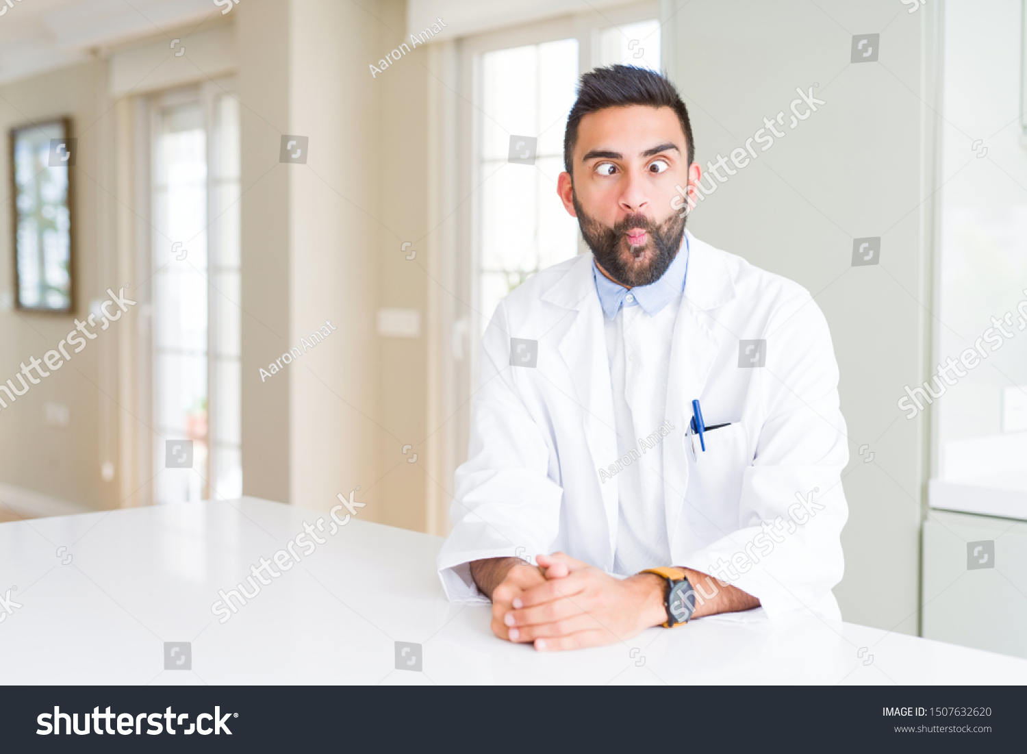 Handsome hispanic doctor or therapist man wearing medical coat at the clinic making fish face with lips, crazy and comical gesture. Funny expression. #1507632620