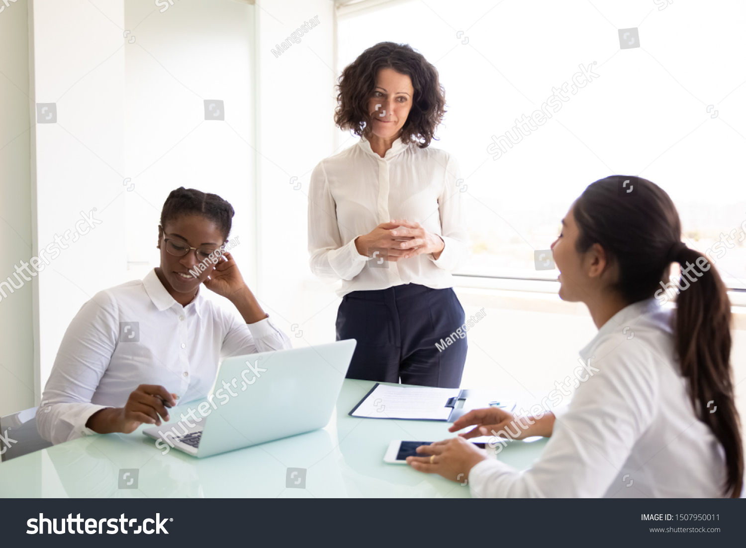 Confident business leader discussing project with team. Businesswomen sitting and standing at conference table, using laptop and talking. Teamwork or leadership concept #1507950011