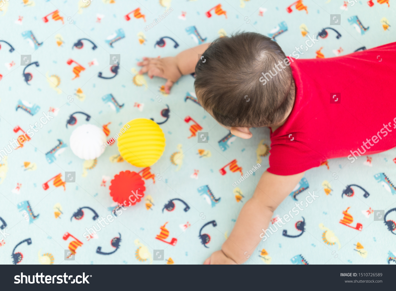 Baby playing with colorful toy rubber balls in the crib in a bright room. Child wearing a red bodysuit, laying on playful dinosaur crib sheet. #1510726589