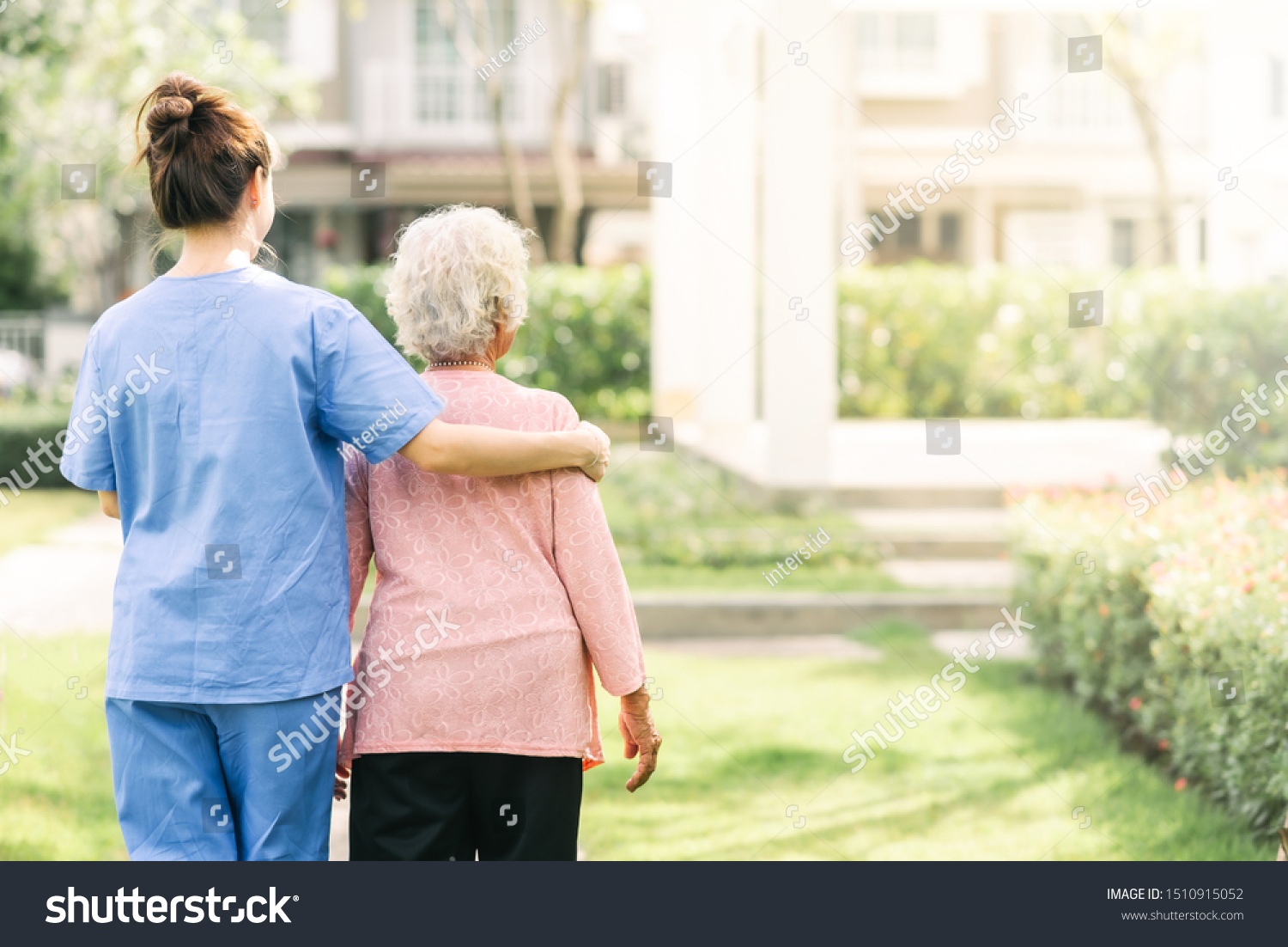 Back view of nurse caregiver support walking with elderly woman outdoor #1510915052