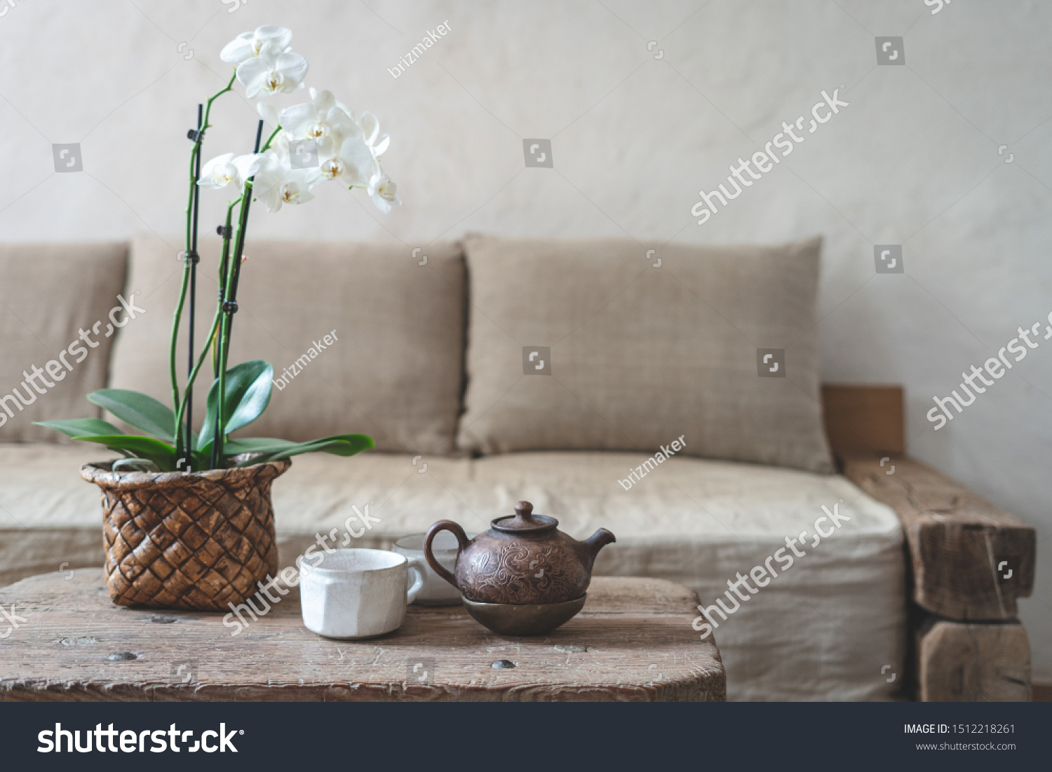 Selective focus of clay teapot, white cup and orchid flower at wooden table against blurred wall with copy space on background. Cozy house with comfort sofa in vintage interior style #1512218261