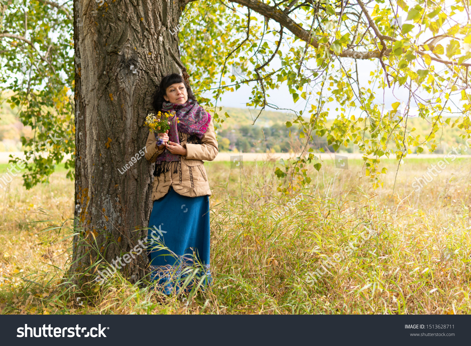 Beautiful woman standing under a tree in a Park in nature. She holds flowers in her hands. Autumn. #1513628711