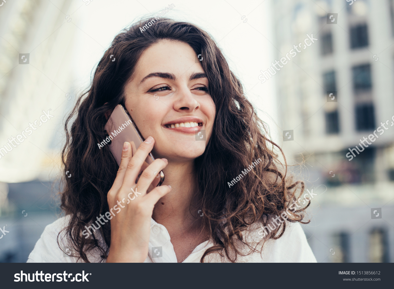 Cheerful pretty young woman talking on phone and look straight ahead. Holding smartphone close to ear. Standing alone outside. White buildings behind. Daylight #1513856612
