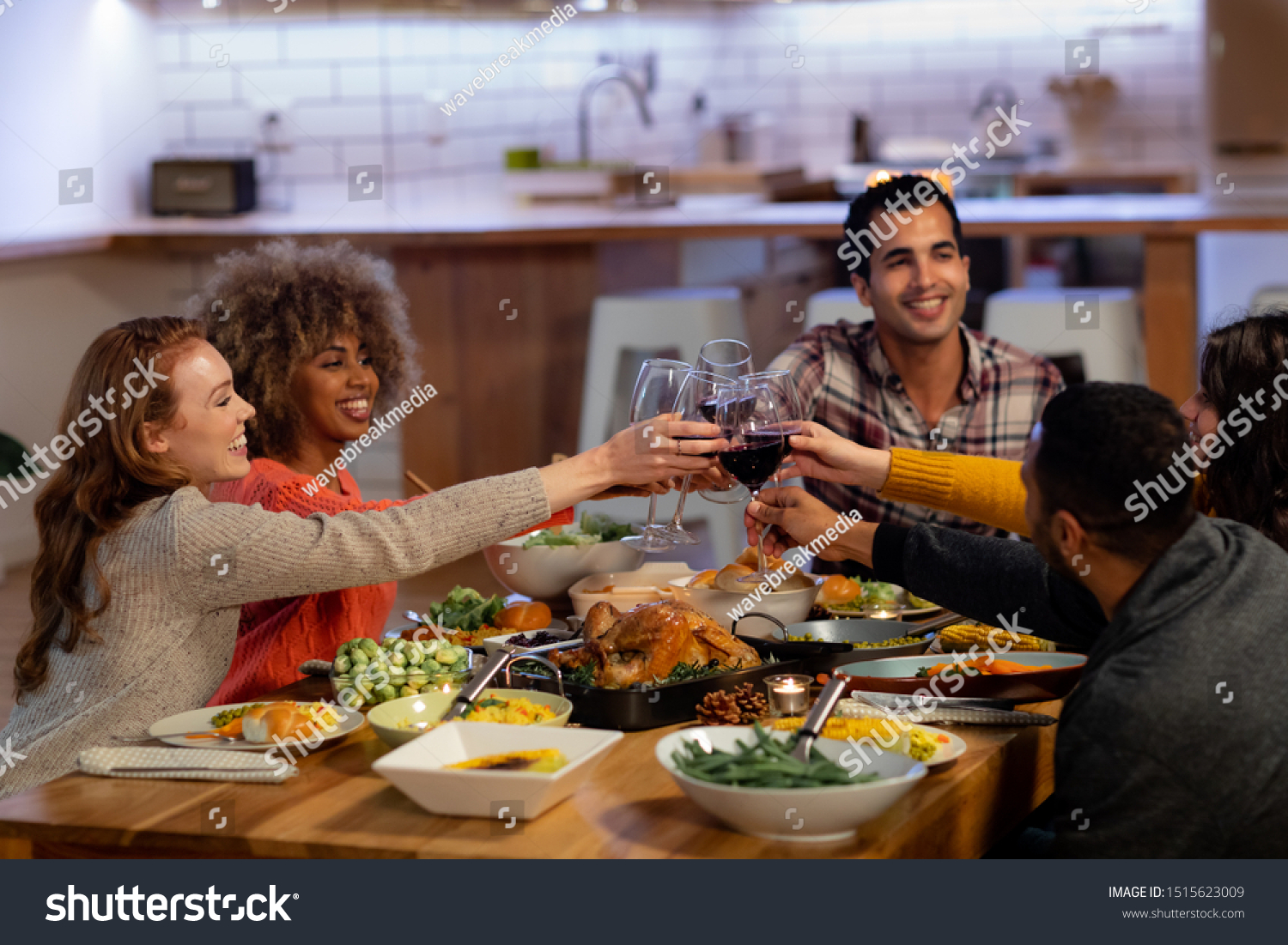 Side view of a group of young adult multi-ethnic male and female friends sitting at a table at home set for Thanksgiving dinner making a toast with glasses of red wine #1515623009