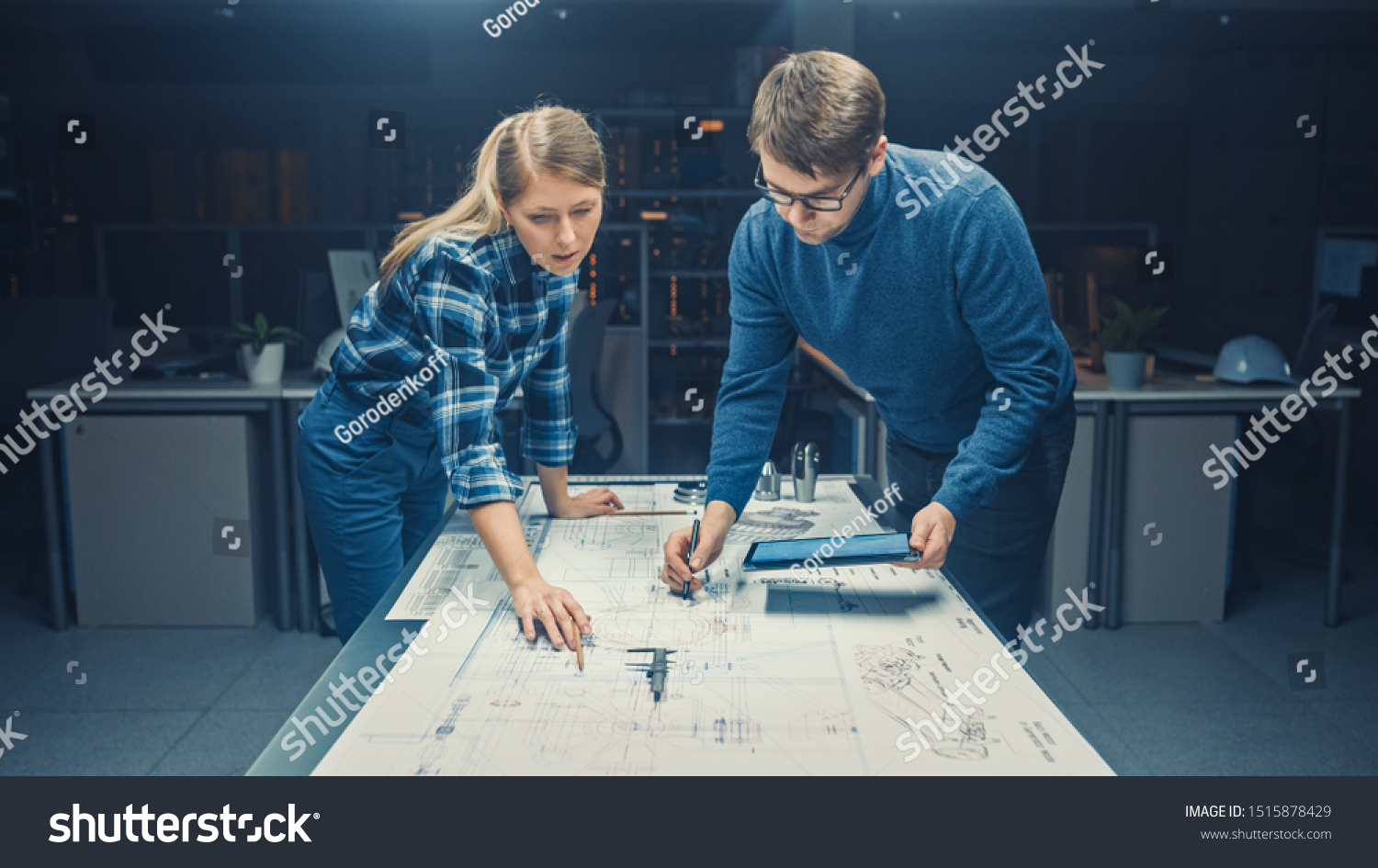 In the Dark Industrial Design Engineering Facility Male and Female Engineers Talk and Work on a Blueprints Using Conference Table. On the Desktop Drawings, Drafts and Electric Engine Components, Parts #1515878429