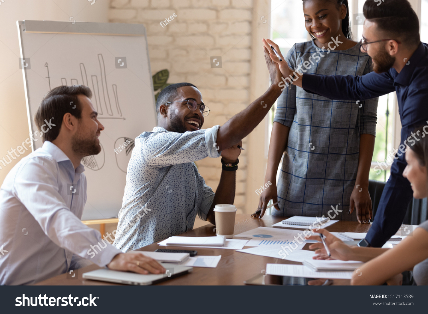 Happy african american male manager sitting at table on business team brainstorming meeting, giving high five to arabian colleague, congratulating with shared teamwork success or good project idea. #1517113589