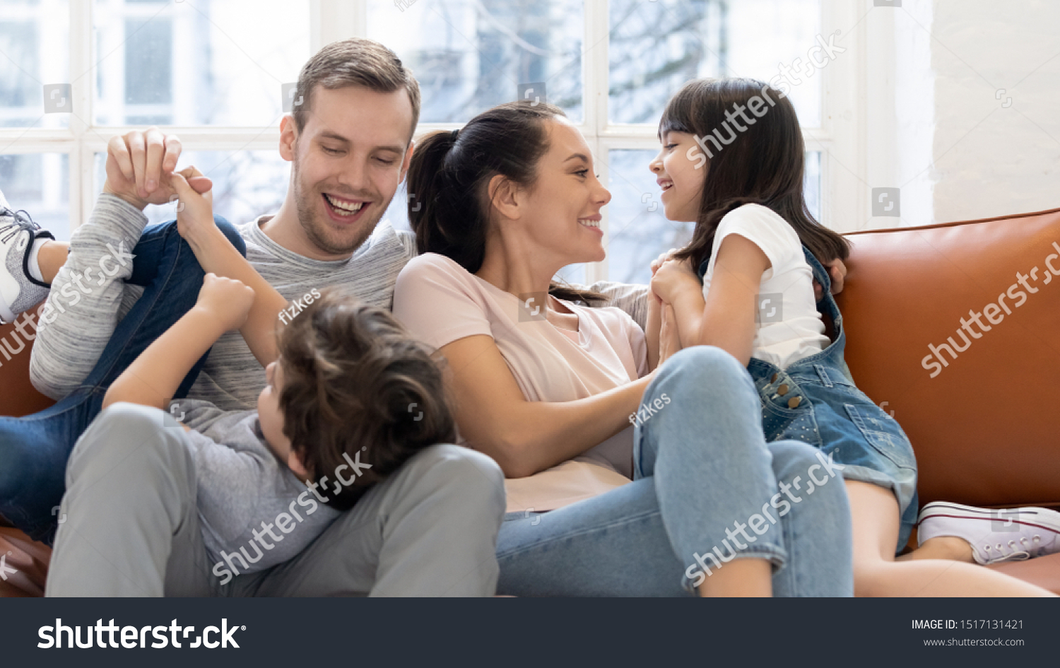 Cheerful family enjoying leisure weekend time together at home. Happy parents mommy and daddy playing with joyful small daughter and son, tickling each other, having fun on couch in living room. #1517131421