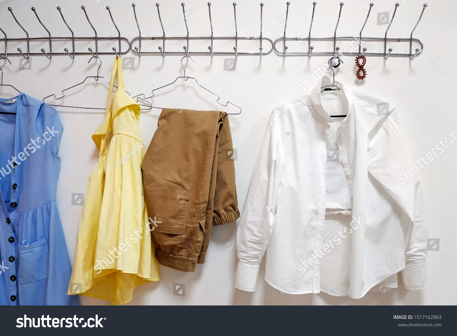 Metal hanger on a white wall. On a hanger are four trempels with clothes: a blue sundress, brown trousers, a white shirt, a yellow dress.  Multi-colored women's clothing, summer wardrobe #1517162963