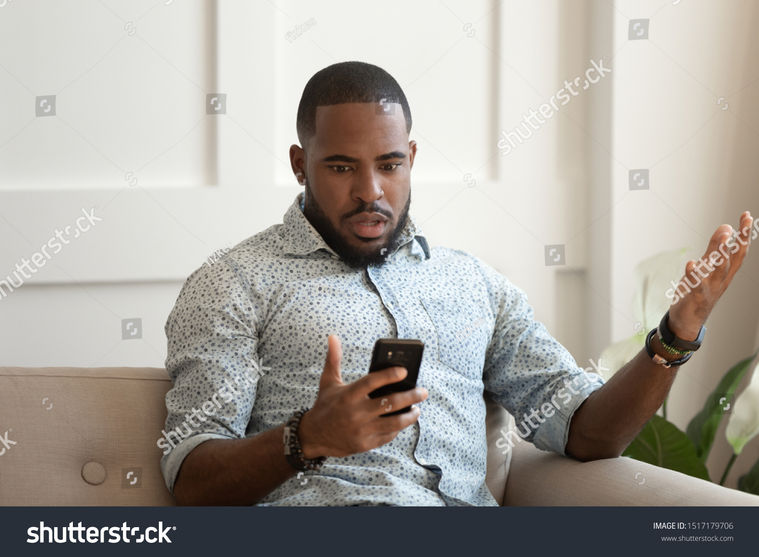 African astonished man sitting on sofa holds cellphone read e-mail sms feels shocked received terrified news, guy looks at online calendar forgot missed important meeting, phone crash problems concept #1517179706