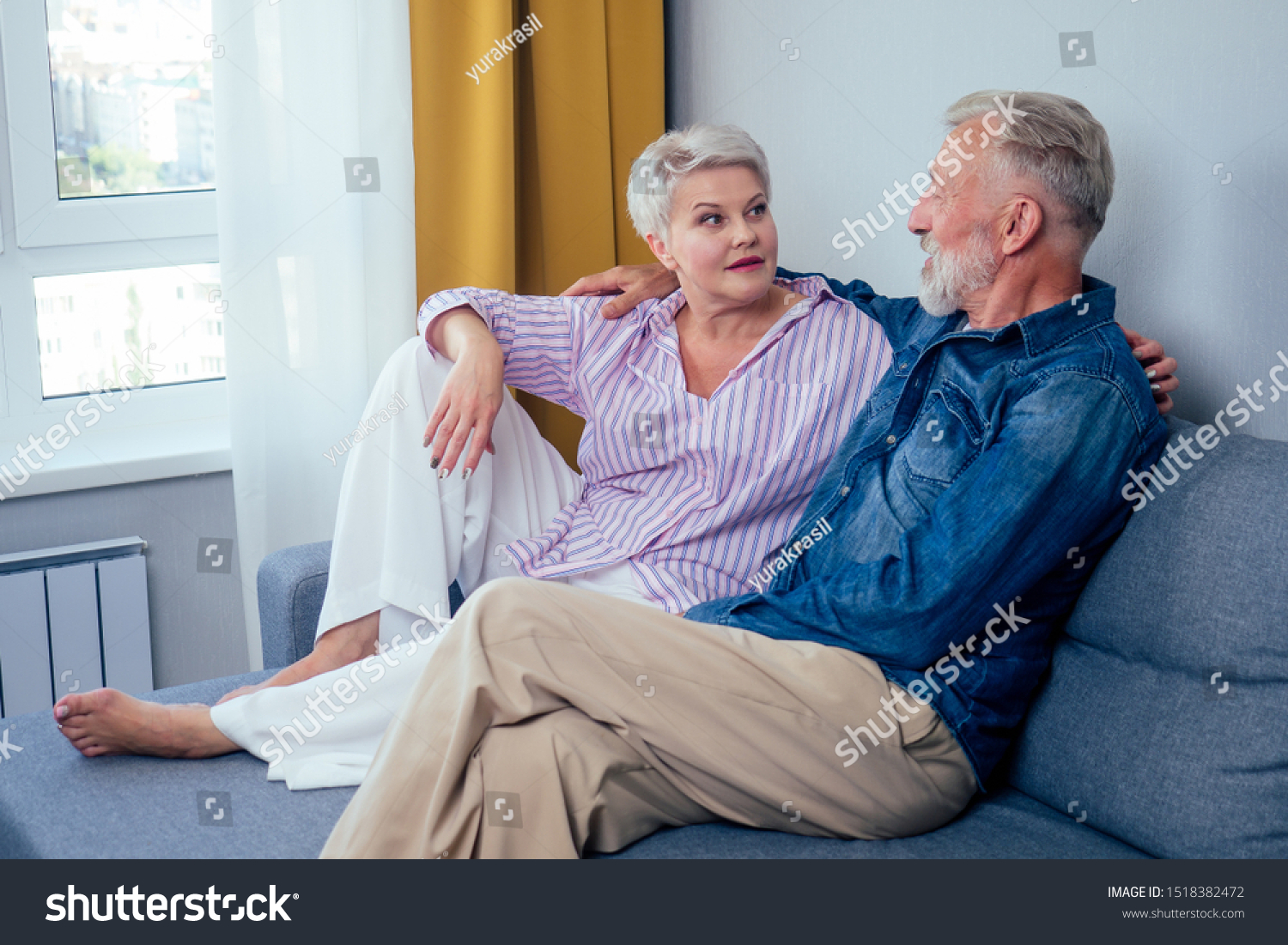 Mature good-looking well dressed couple sitting in sofa in day light from big window and yellow curtains in living room,touching hand . #1518382472