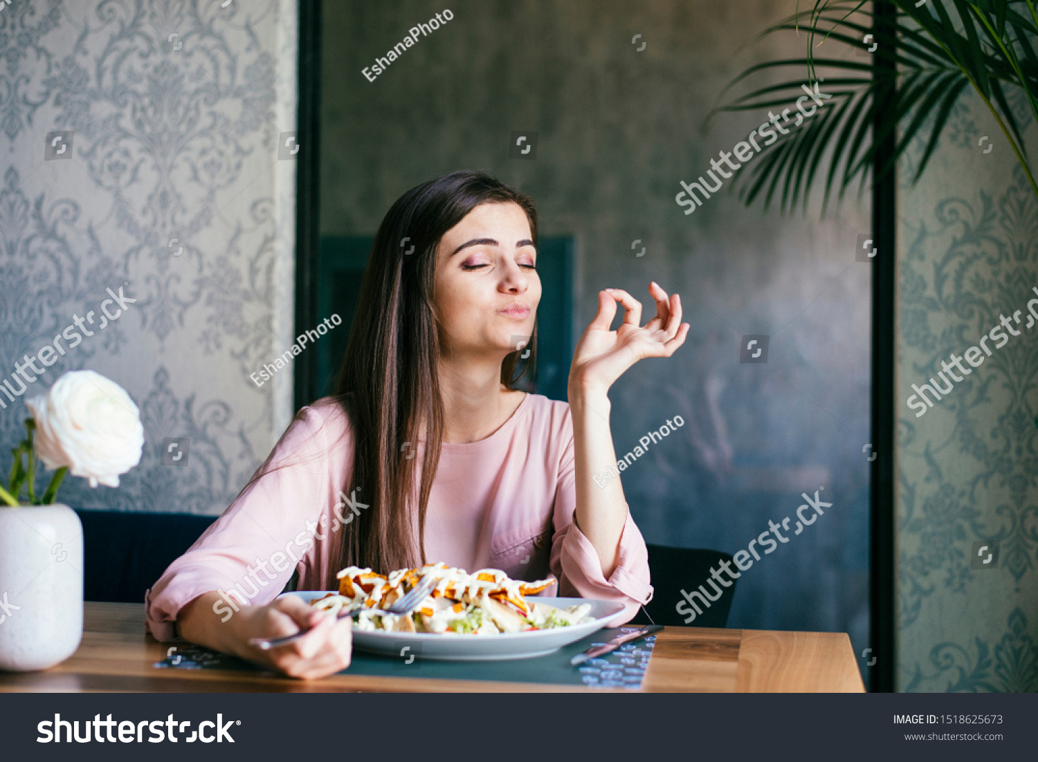Pretty young lady sitting in a beautiful restaurant, enjoying lunch or dinner and making delicious hand gesture to express how good the food is.  #1518625673