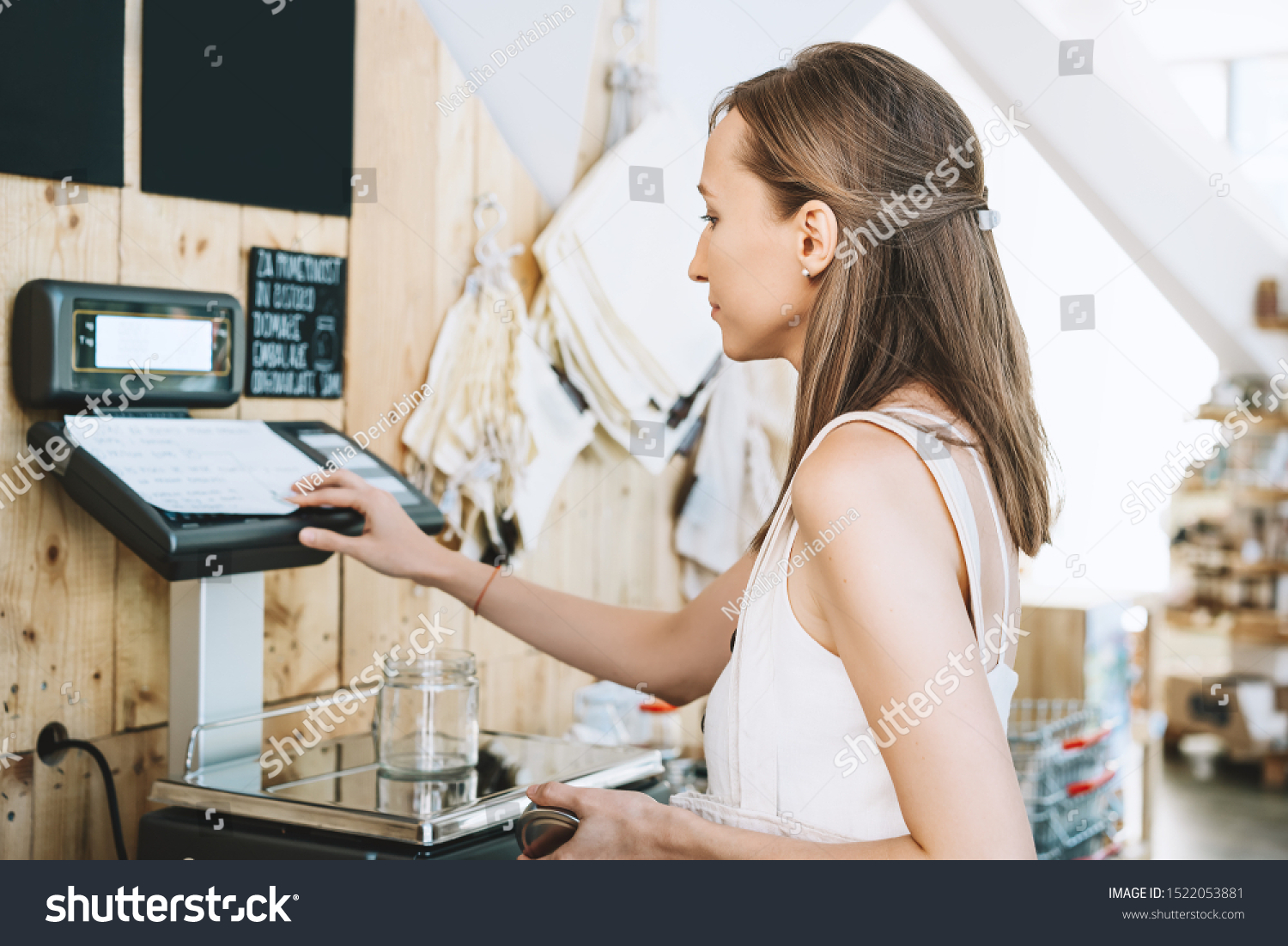 Woman chooses and buys products in zero waste shop. Weighing dry goods in plastic free grocery store. Girl with cotton reusable bag weigh glass jars on scales. Eco shopping at local business #1522053881