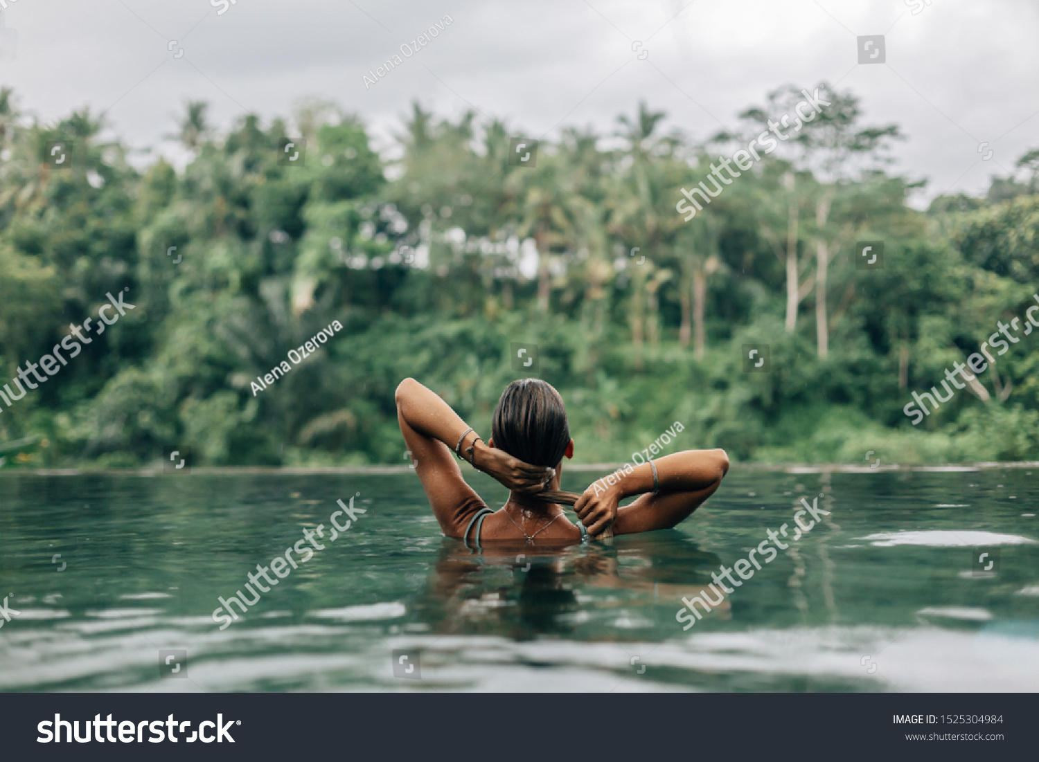 Happy woman enjoying warm tropical rain falling on her while swimming in infinity pool with a jungle view in Ubud, Bali #1525304984