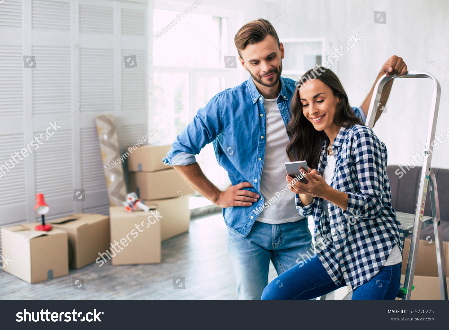 Checking for ideas. Handsome man in denim outfit is checking something in the smartphone together with his gorgeous wife, leaning on the ladder with his left hand. House moving concept. #1525770275